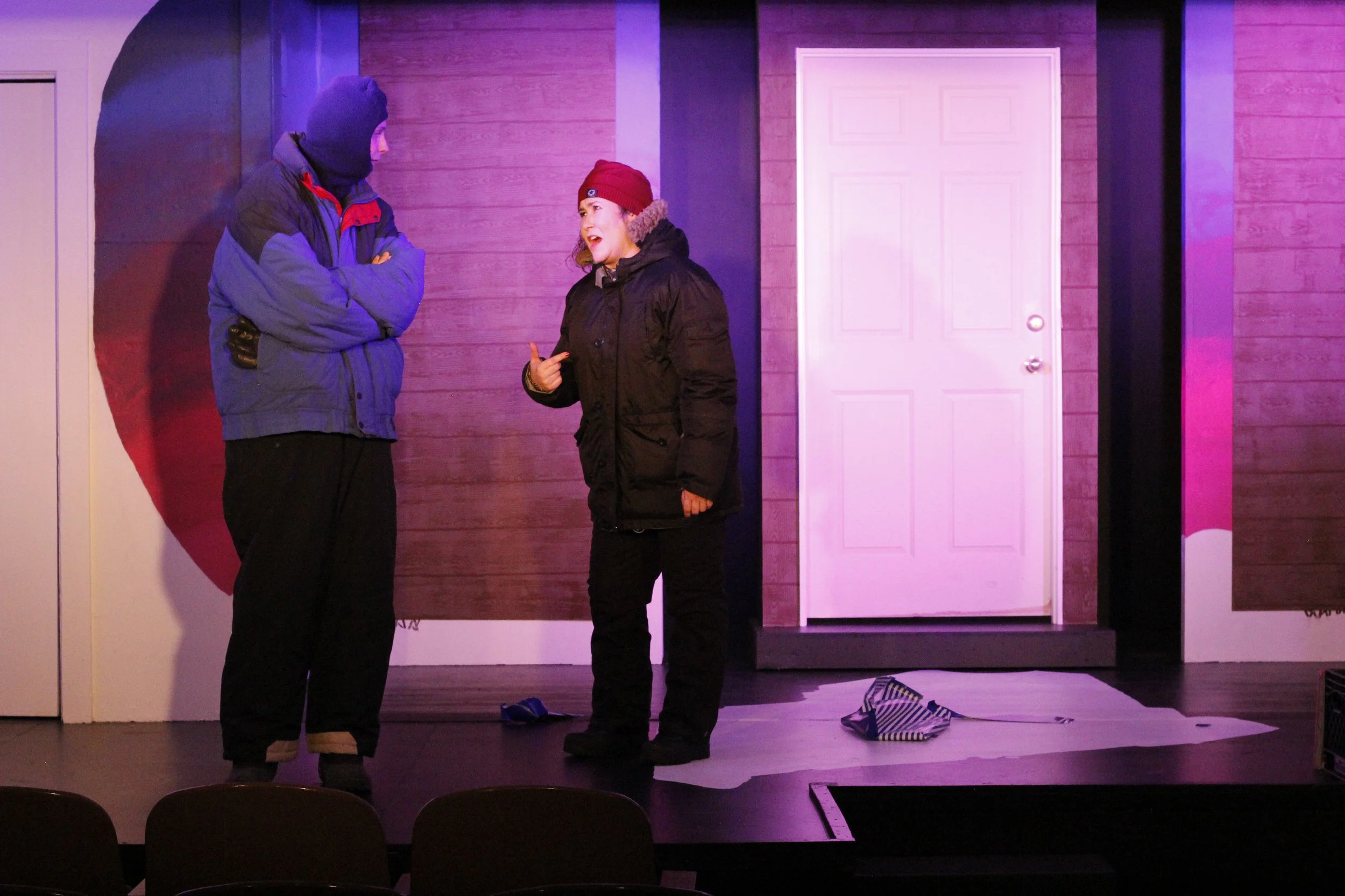 Two actors on stage during a theatrical performance, dressed in winter clothing, having a conversation with a purple backdrop and door in the background.
