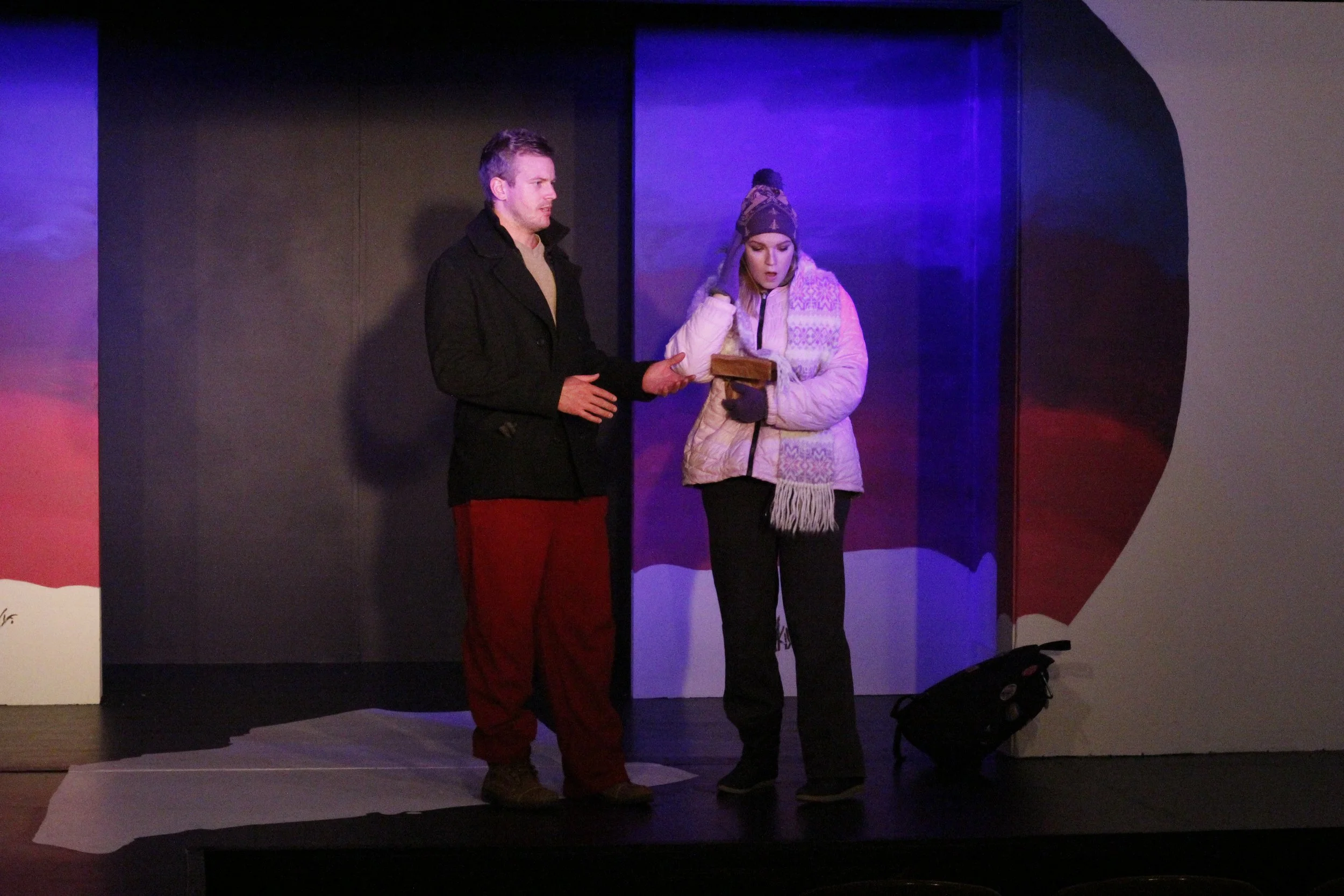 Two actors on a stage, one man and one woman, engaged in a scene. The man is wearing a black coat and red pants, and the woman is in a gray jacket with a knit hat and scarf, holding a small box. The background features a dark, painted backdrop with u