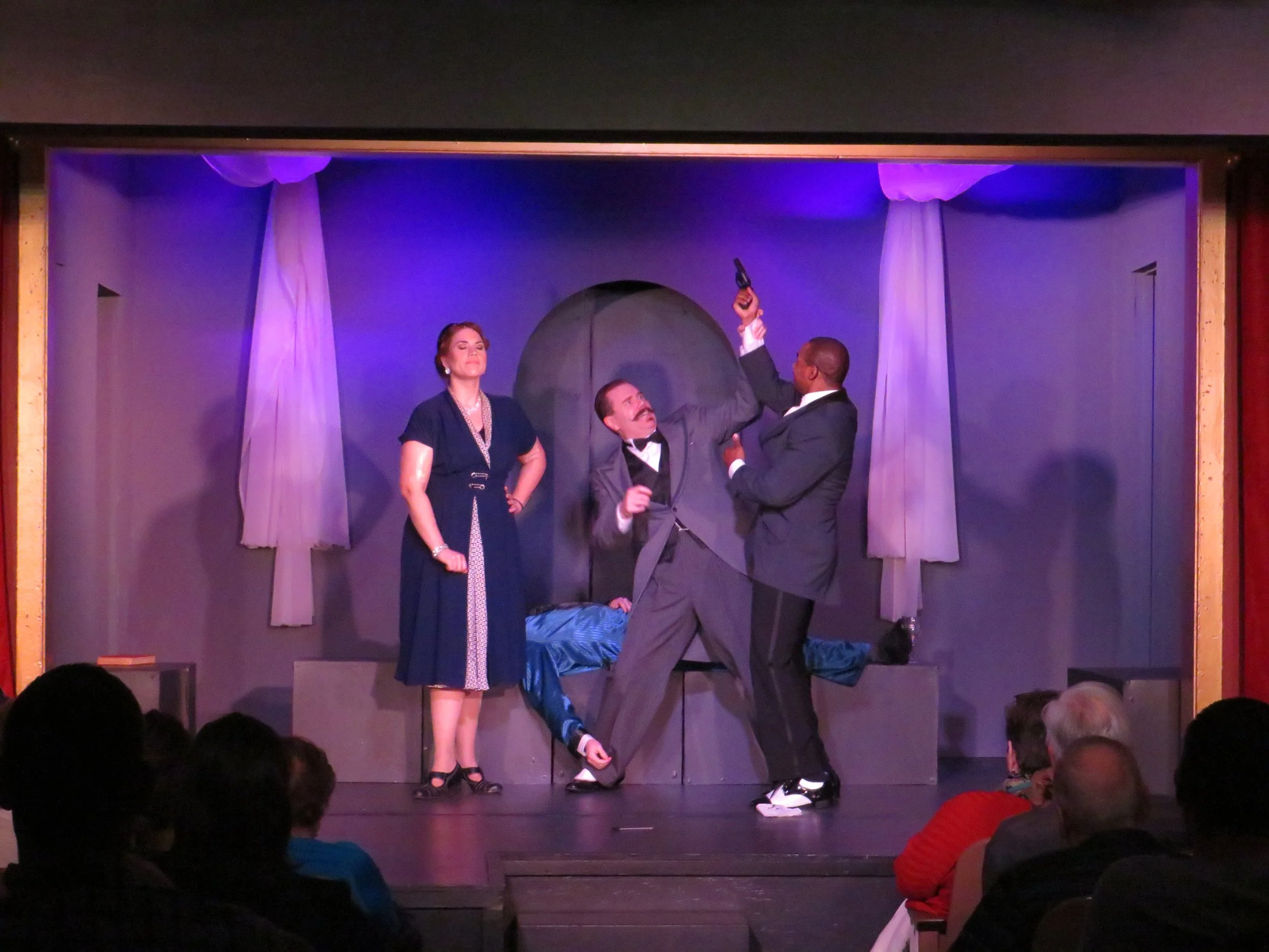 Actors performing a scene on stage with purple lighting, with three people in formal attire; one man is on the ground, another man is holding a gun, and a woman stands to the side watching.