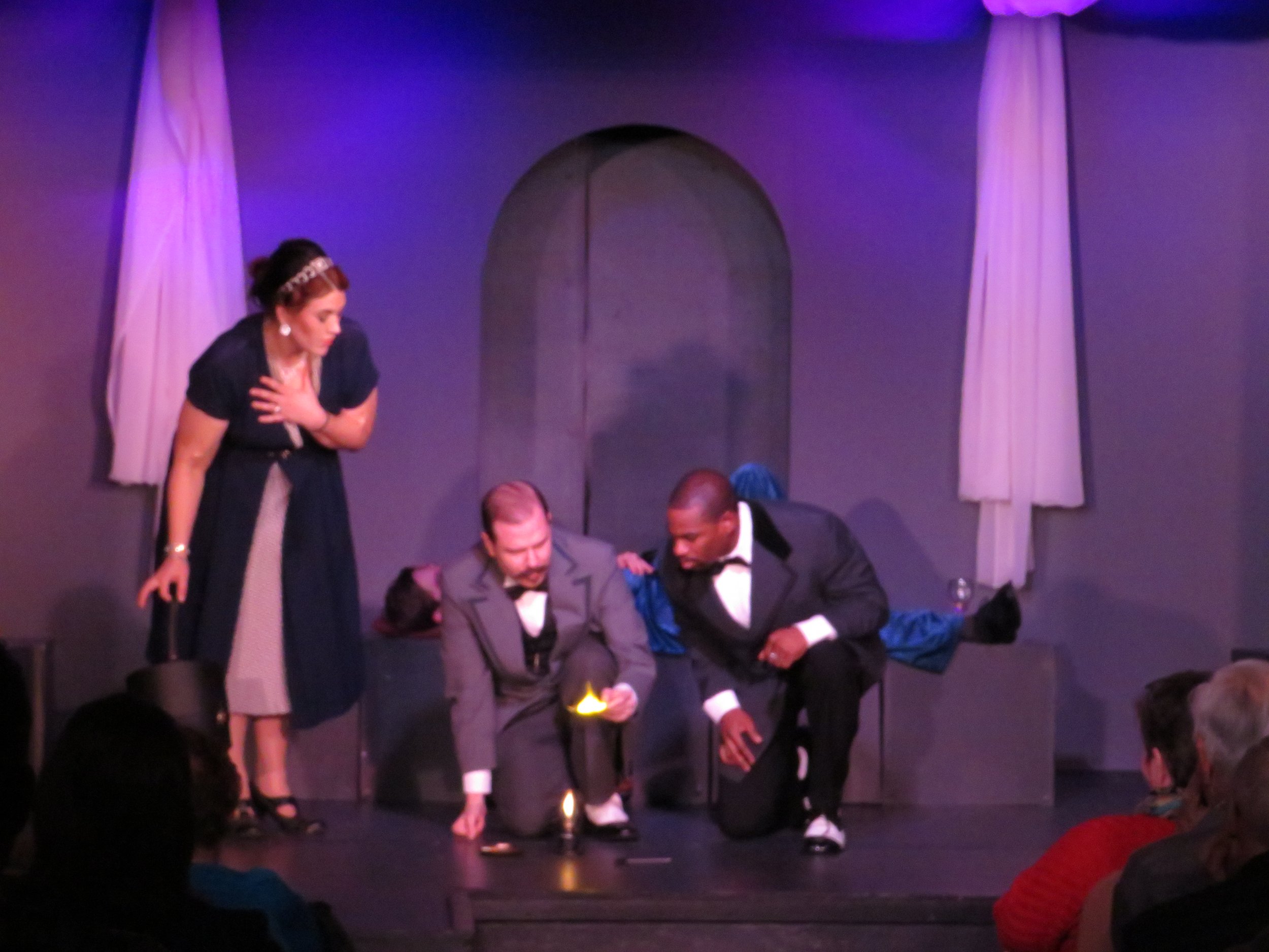 Actors performing on stage with purple lighting, with one man lying down, two men kneeling examining an object with light, and a woman standing nearby