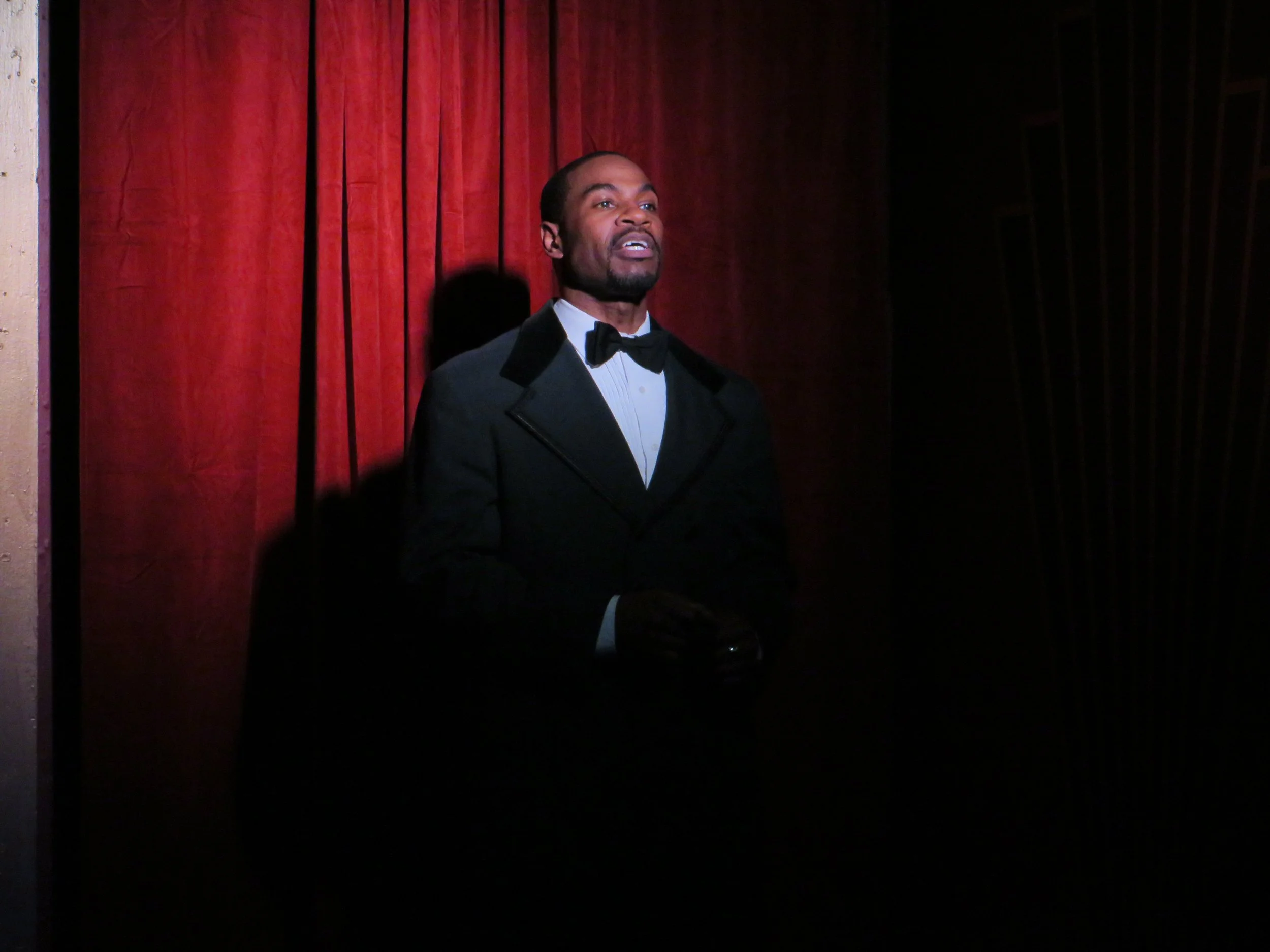 A man dressed in a tuxedo standing in front of red velvet curtains, illuminated by a spotlight.