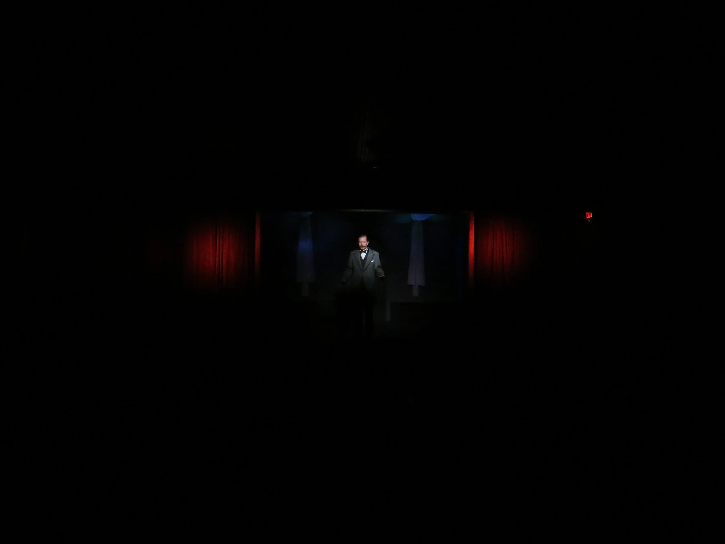 A man in a tuxedo standing on a dark stage with red curtains and a black backdrop, illuminated by a spotlight.