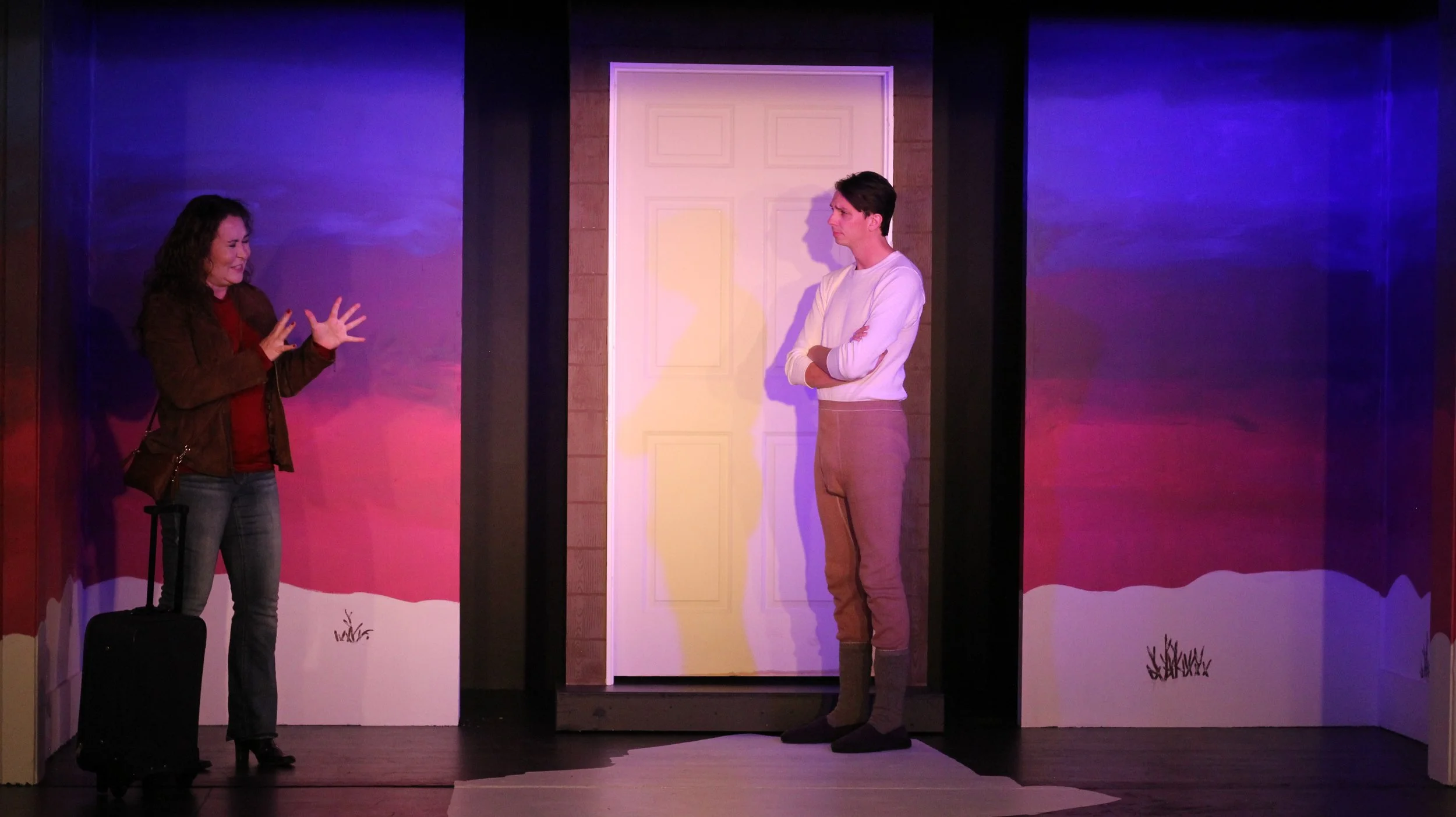 A woman and a man are performing a scene on stage; the woman has curly dark hair, is gesturing with her hands, and has a suitcase nearby, while the man with dark hair stands with his arms crossed in front of a white door.
