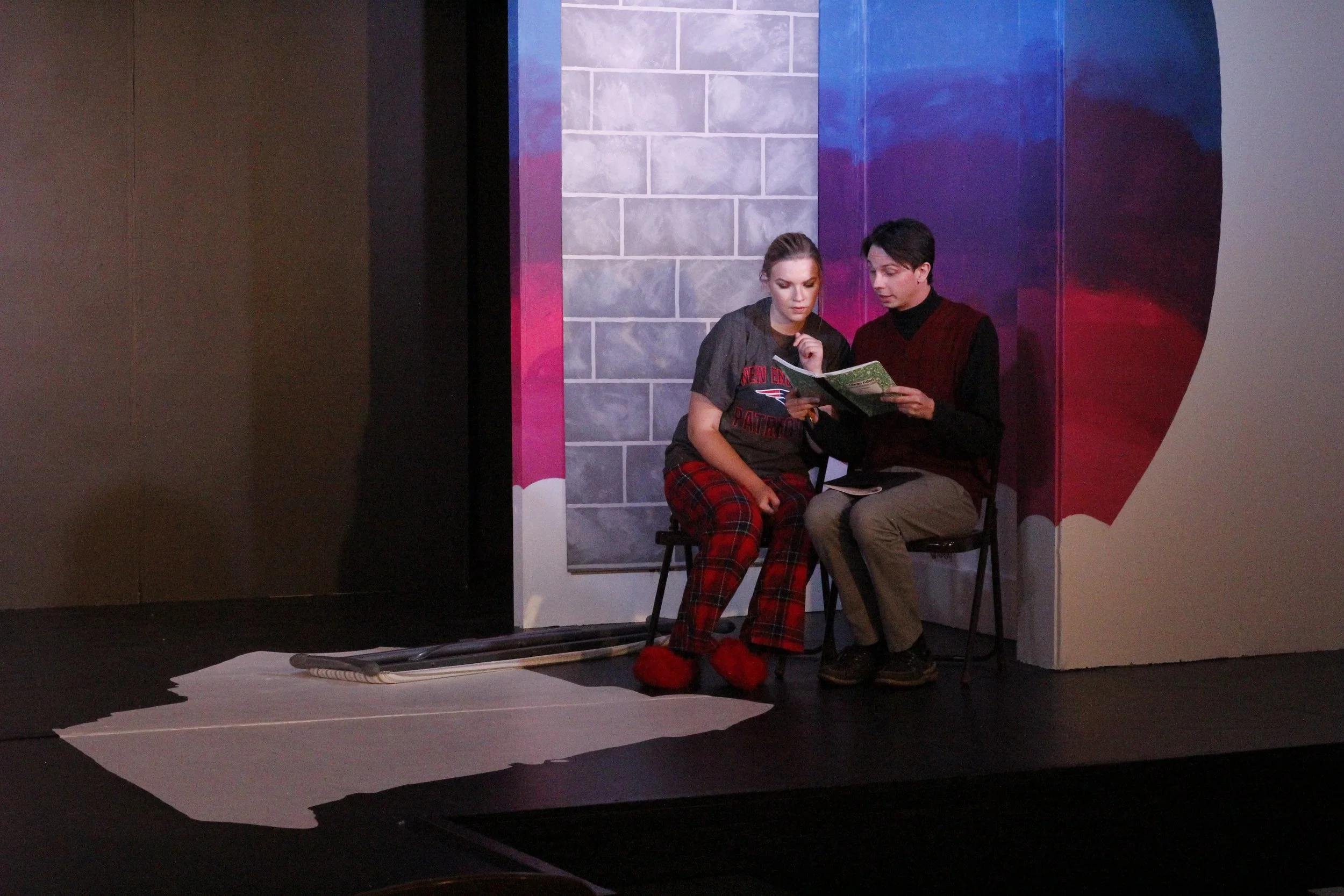 Two young people sitting on chairs on a stage, reading a book together, with a painted background and some fabric and cords on the floor in front of them.
