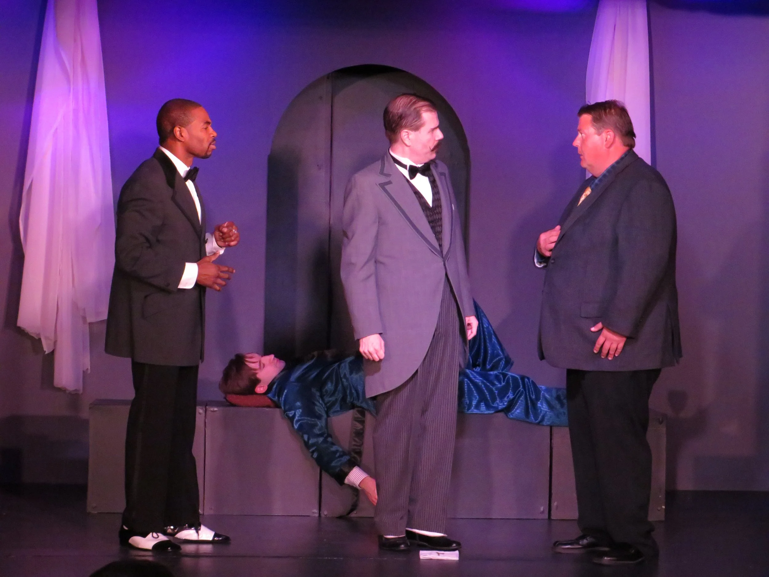 Four men on stage, three standing and one lying down, in a scene from a theatrical play. The man in the center wears a gray suit with a black vest, the man on the right uses a checkered blazer, and the man on the left is in a tuxedo. The man lying do