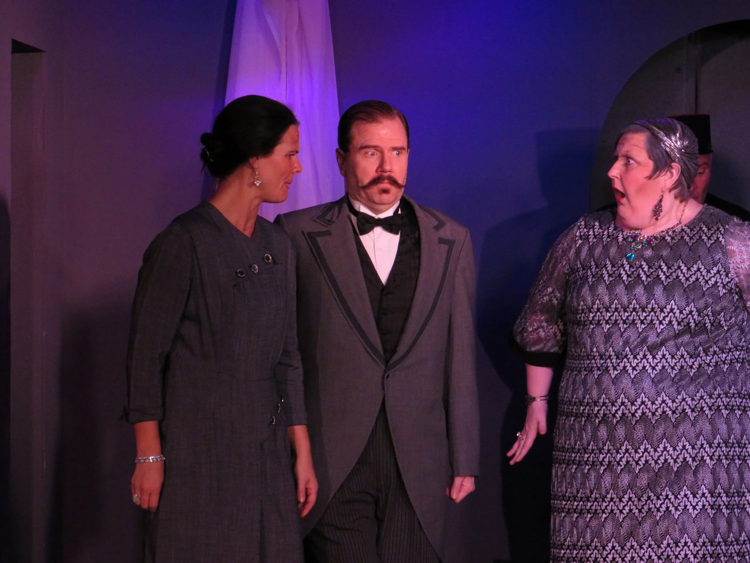 Three actors in period costumes engaged in a dramatic scene, woman on left with dark hair in bun, man in middle with mustache and tuxedo, woman on right with grey hair and patterned dress, inside a theatrical stage setting.
