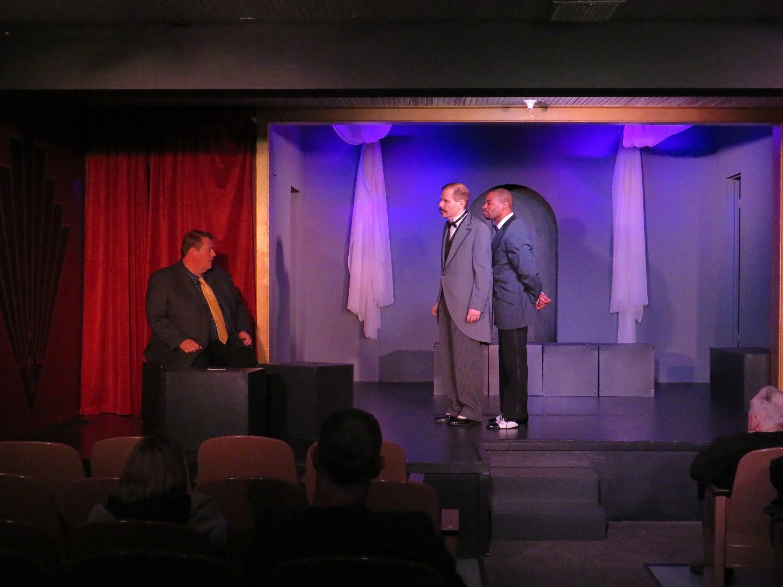 Three men in formal attire on stage, with two men facing each other and a third man sitting at a desk, in a theater setting with red curtains and blue lighting.