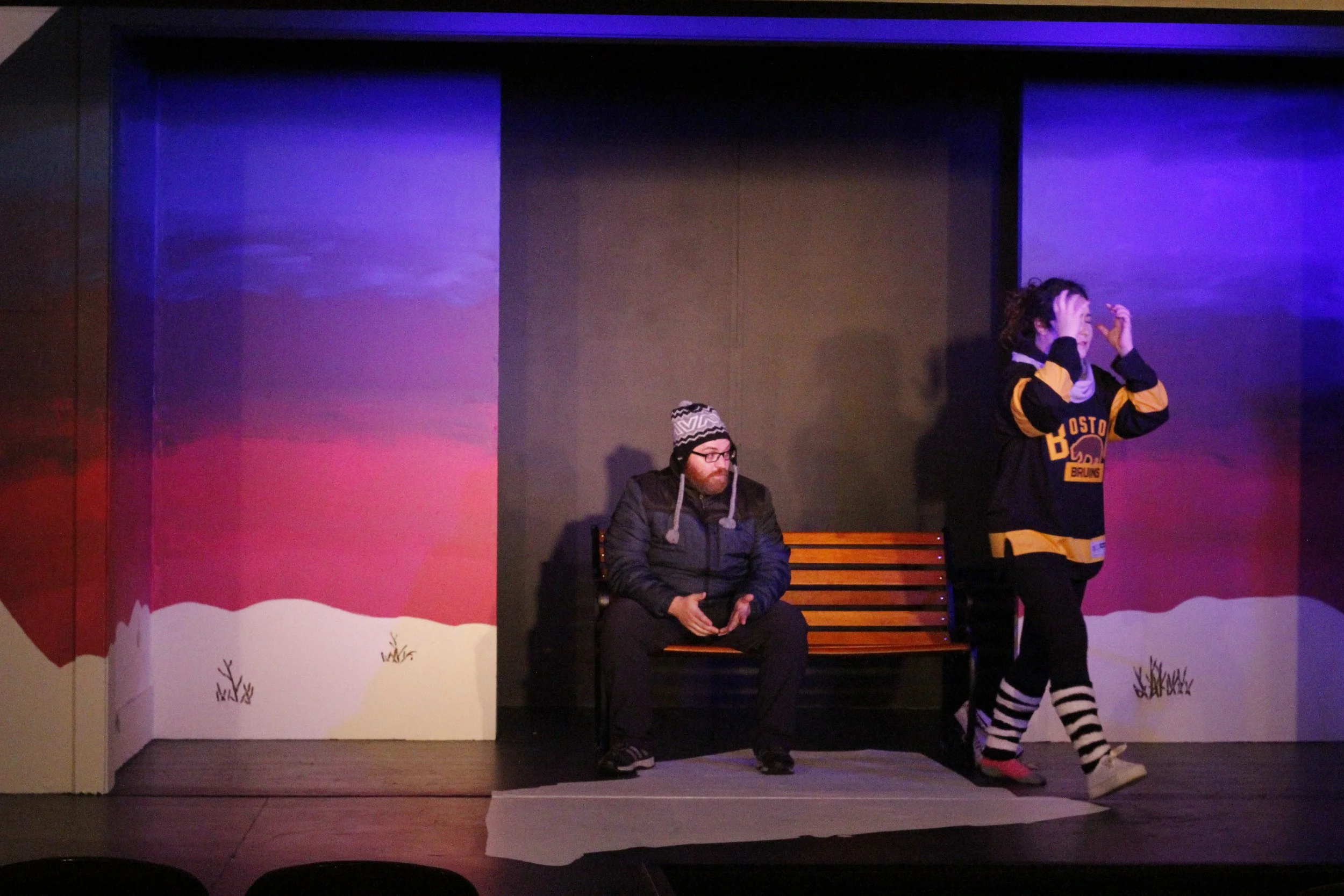 A man sitting on a bench with a winter hat and glasses, looking at his phone, and a woman walking past him on stage, wearing a black and yellow hockey jersey, striped socks, in a theatre with a painted winter scene backdrop.