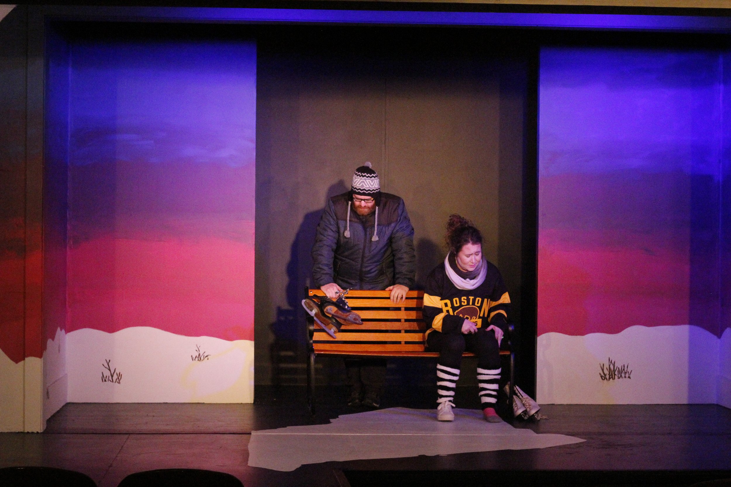 Two people sitting on a wooden bench in a theatrical stage setting with winter scenery painted on the walls, one man standing and leaning on the bench, and one woman sitting with her head bowed.