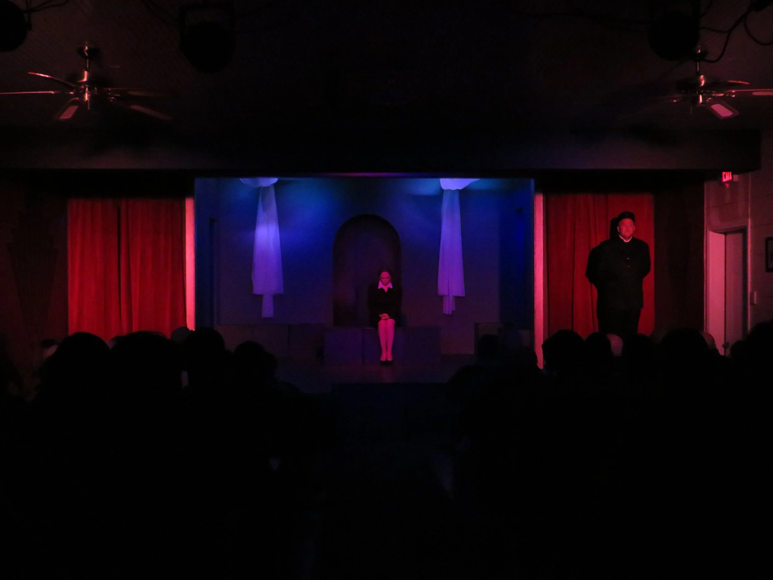 A dimly lit theater stage with a woman sitting on a bench center stage and a man standing to the right. Red curtains are on either side of the stage, and white fabric hangs from the ceiling. Audience visible in the dark foreground.