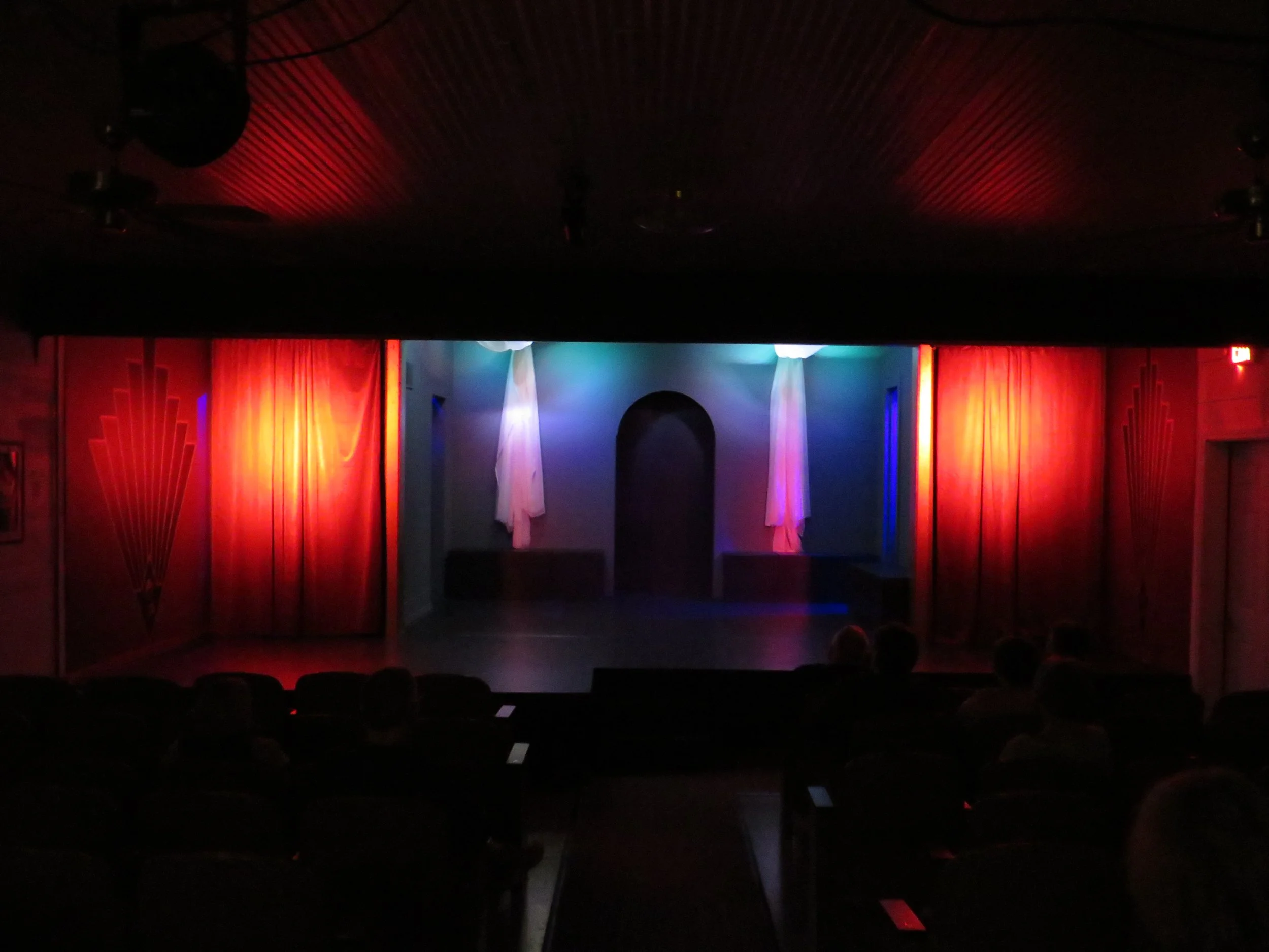 Empty theater stage with red curtains, blue and purple lighting, and an audience seated in the dark.