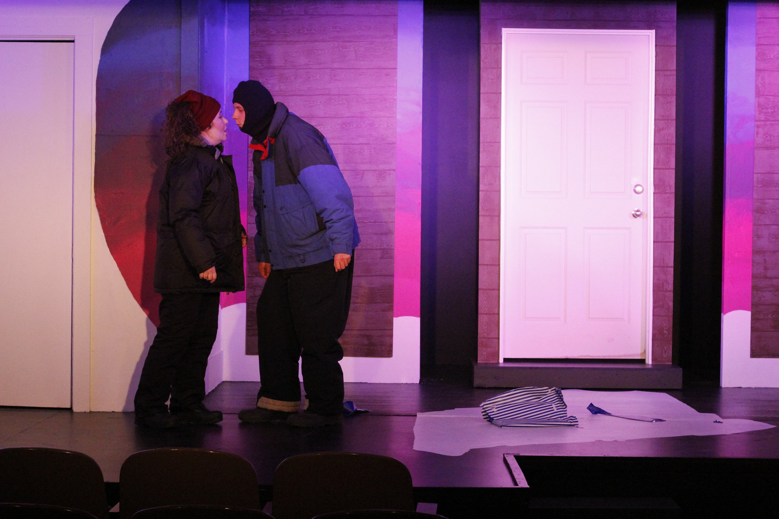 Two actors on stage in a theatrical play, facing each other closely, with a door and set pieces behind them, one dressed in a black jacket and the other in a blue and black jacket, in a scene lit with purple lighting.