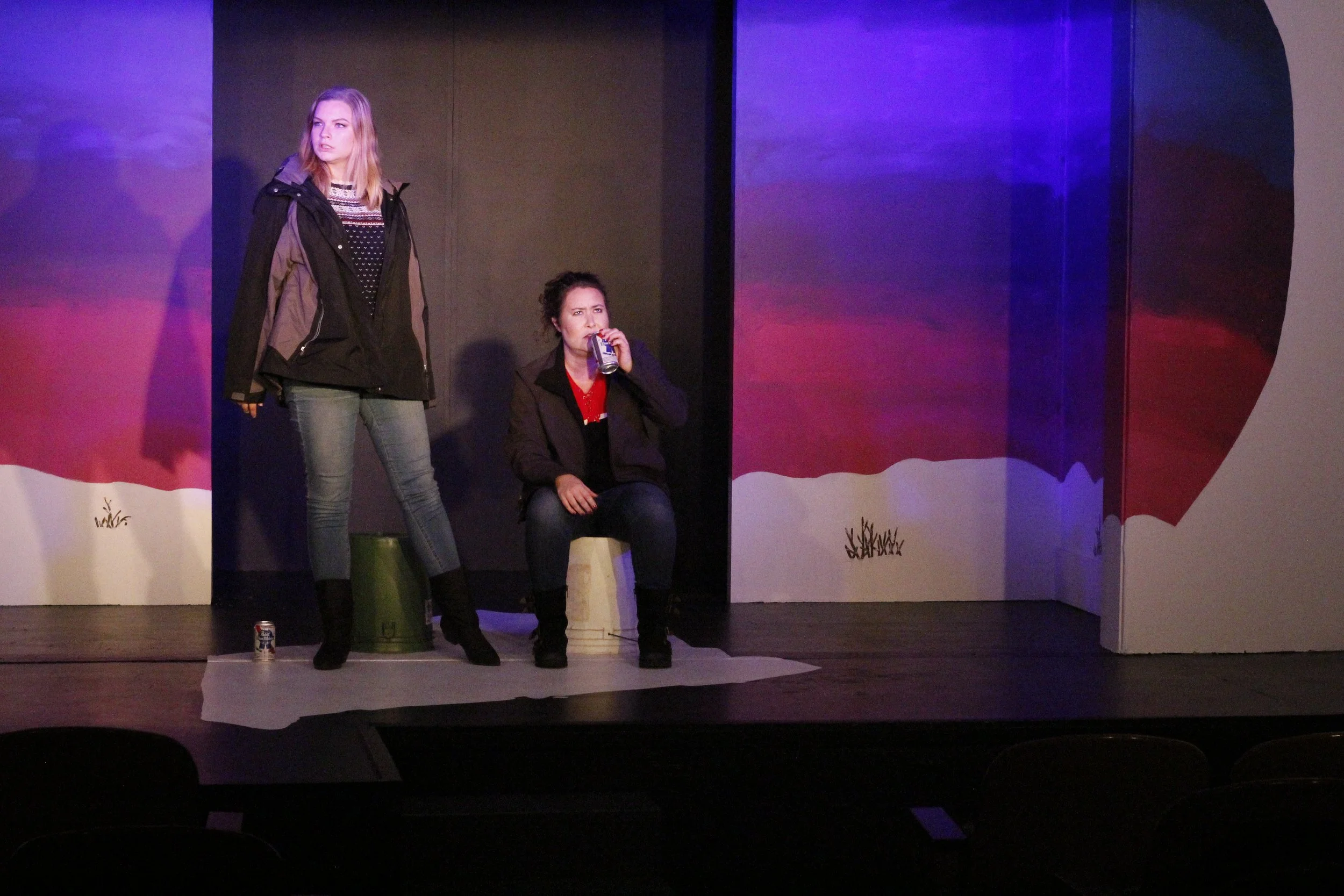 Two women on stage, one standing and one sitting, with painted backdrop and props, during a theatrical performance.
