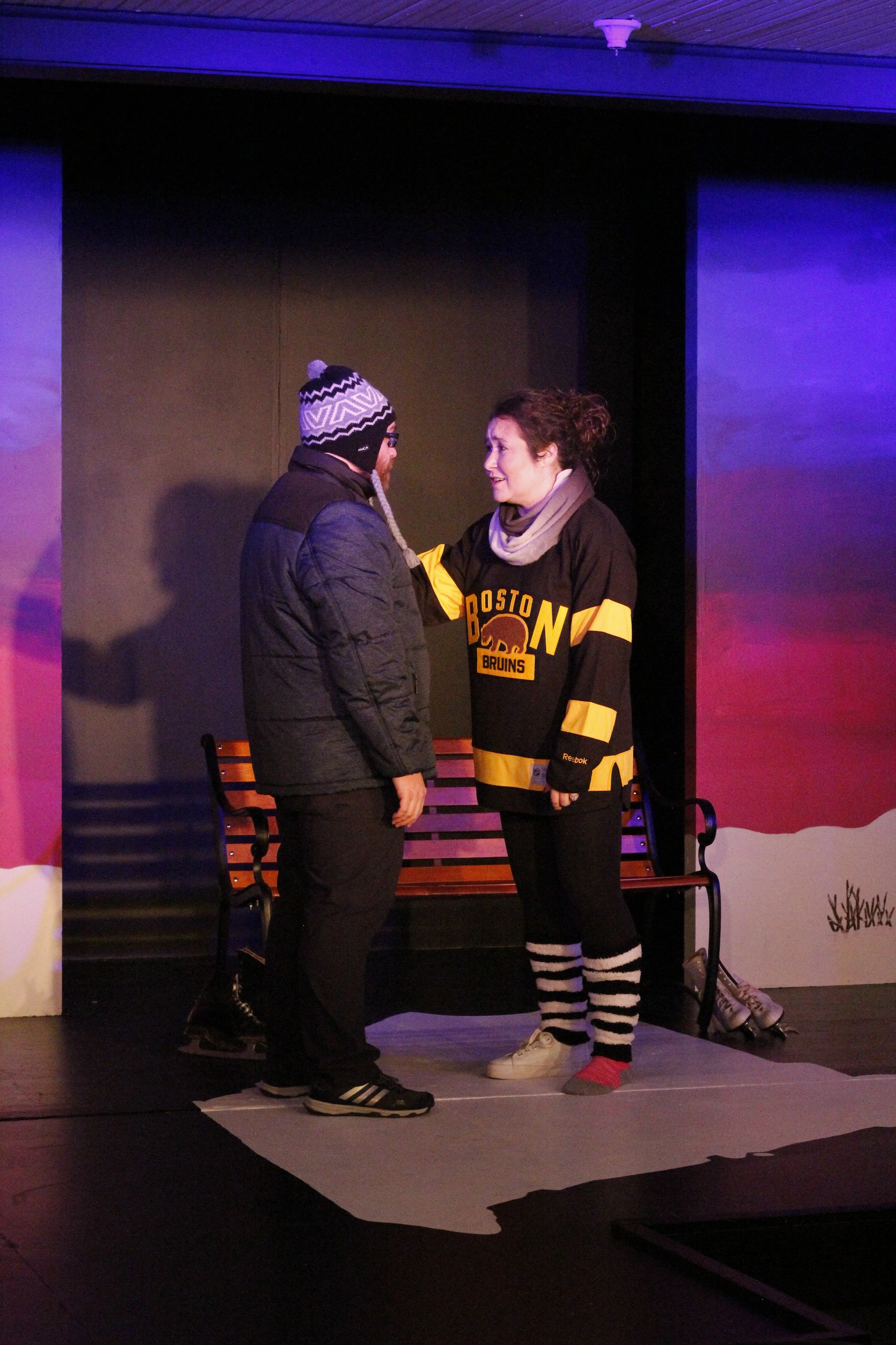 Two people standing on stage, engaged in an emotional conversation, with benches and stage scenery in the background.