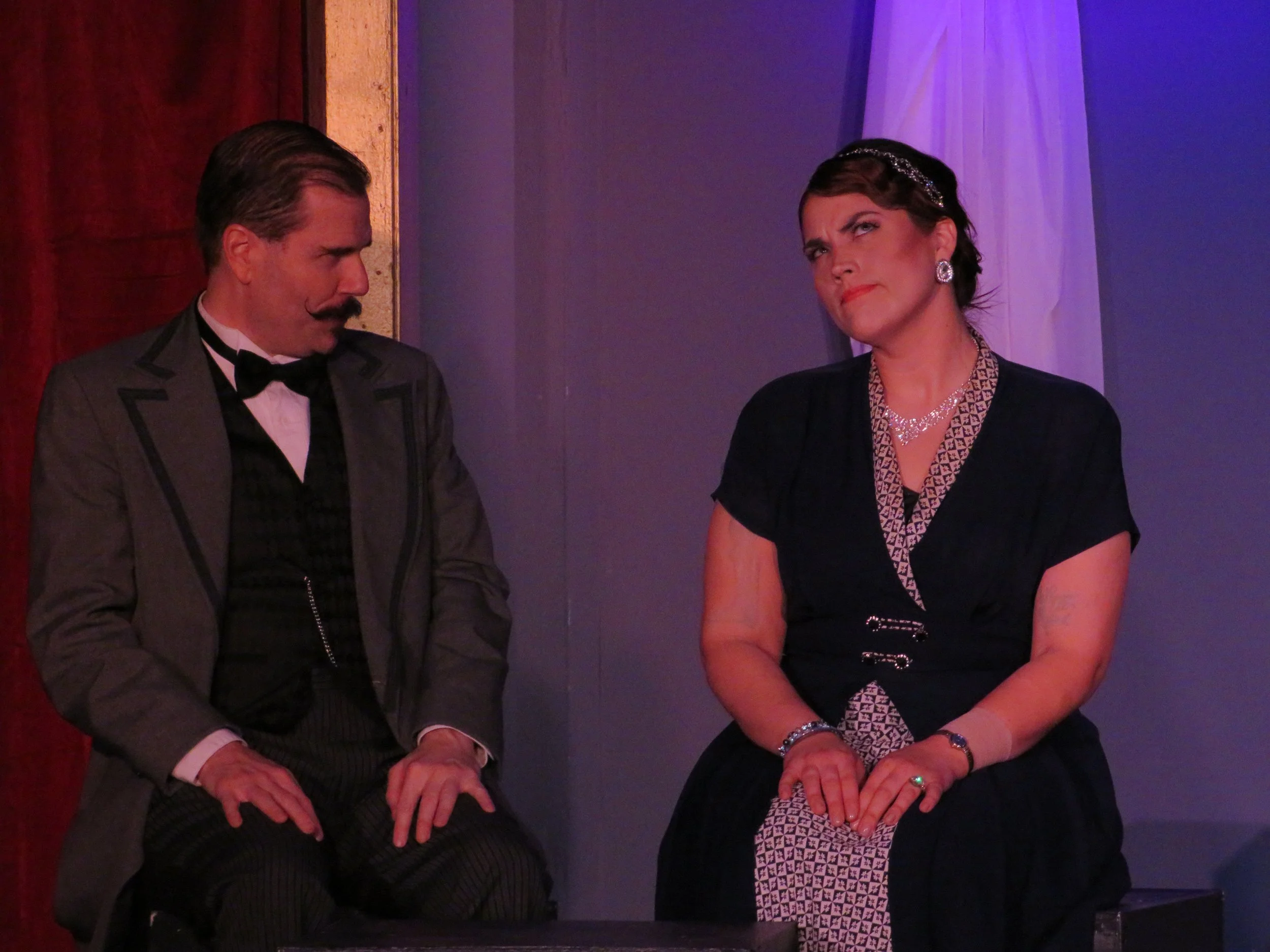 A man and woman sitting next to each other at a formal event, dressed in vintage clothing, with the man looking at the woman and the woman looking slightly upward, both with serious expressions.