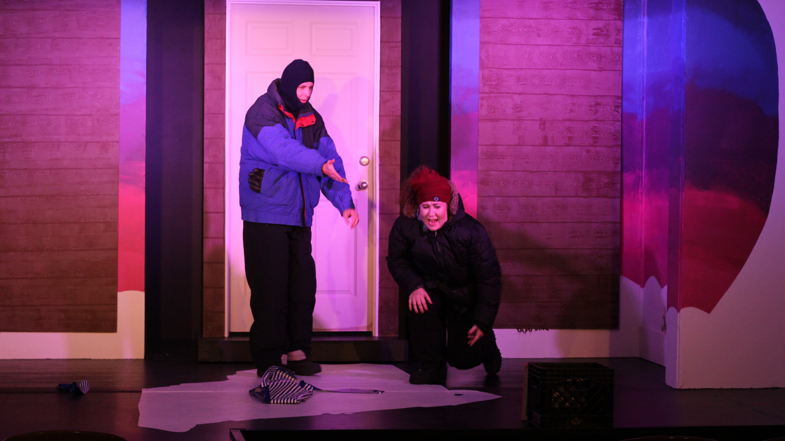 Two performers on a stage with a pink and purple lighting backdrop, one standing and pointing, the other kneeling and appearing distressed, with props including a crate, a striped fabric, and a sock on the floor.