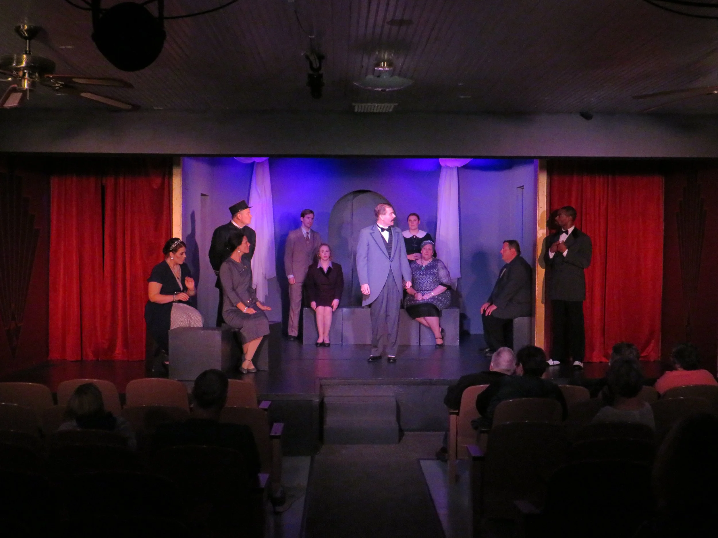 A theatrical play on stage with nine actors, some standing and some seated, in a scene with blue and purple lighting, red curtains on the sides, and audience members watching in front.