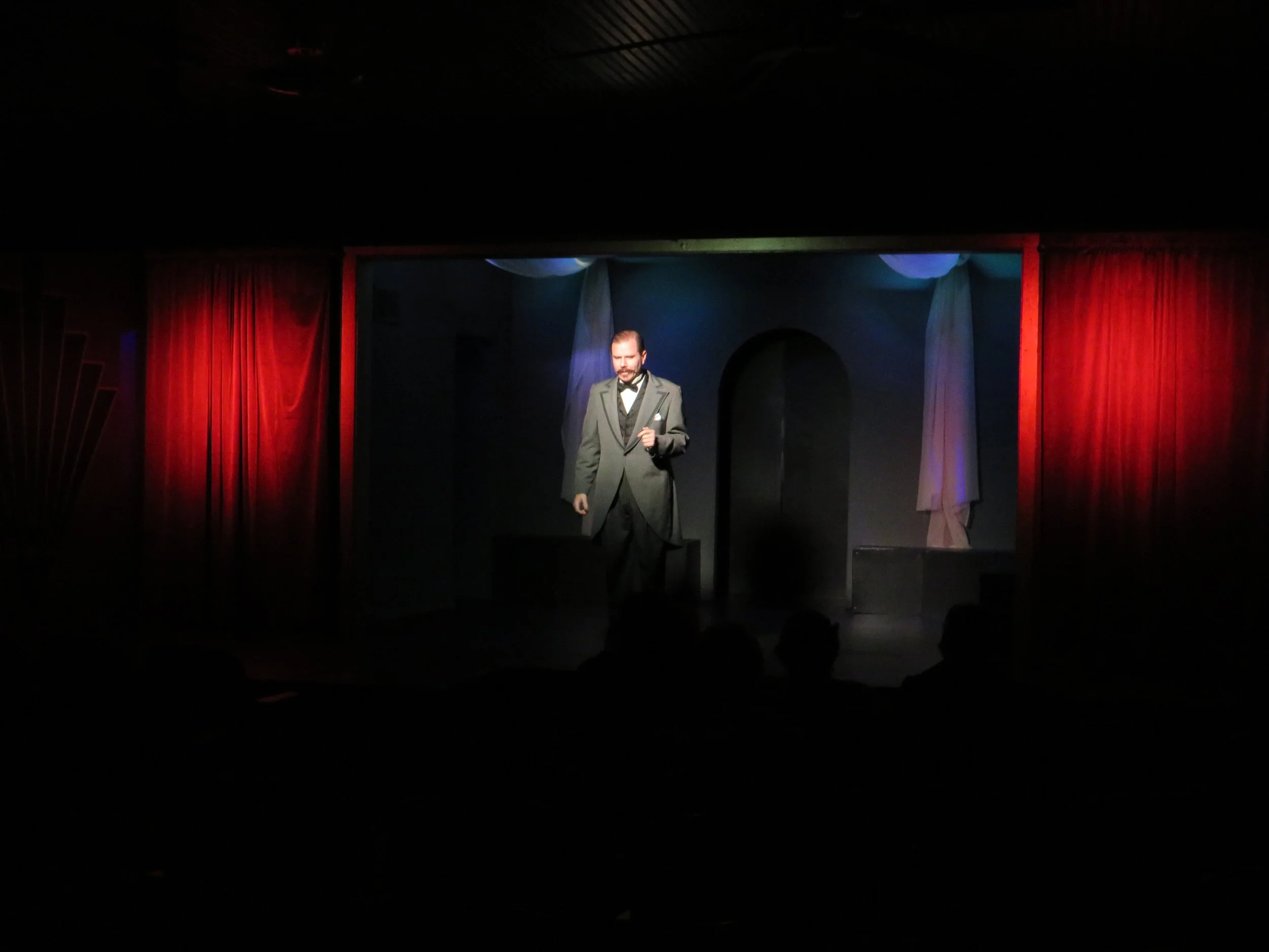 A man in a tuxedo on a stage with red curtains, holding a microphone, performing in a theater.
