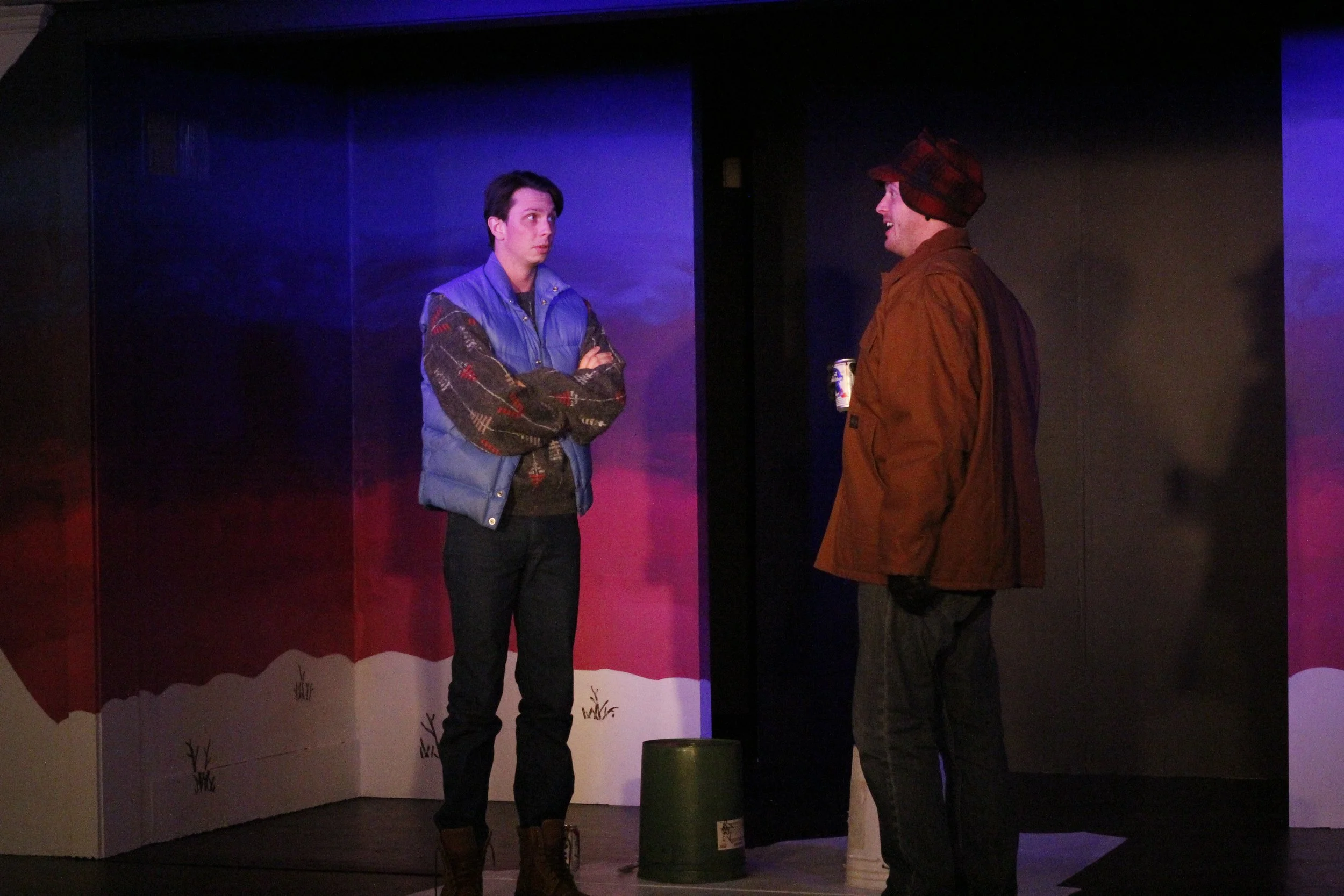 Two men standing and talking on a stage with winter-themed background, one in a blue vest and sweater with arms crossed, the other in a brown jacket and hat holding a cup.