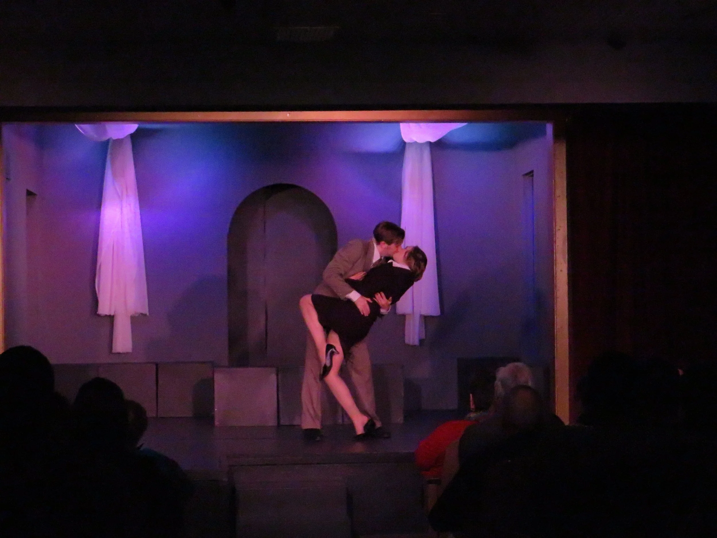 Two people performing a romantic dance or scene on a stage with white curtains and purple lighting, with an audience watching.