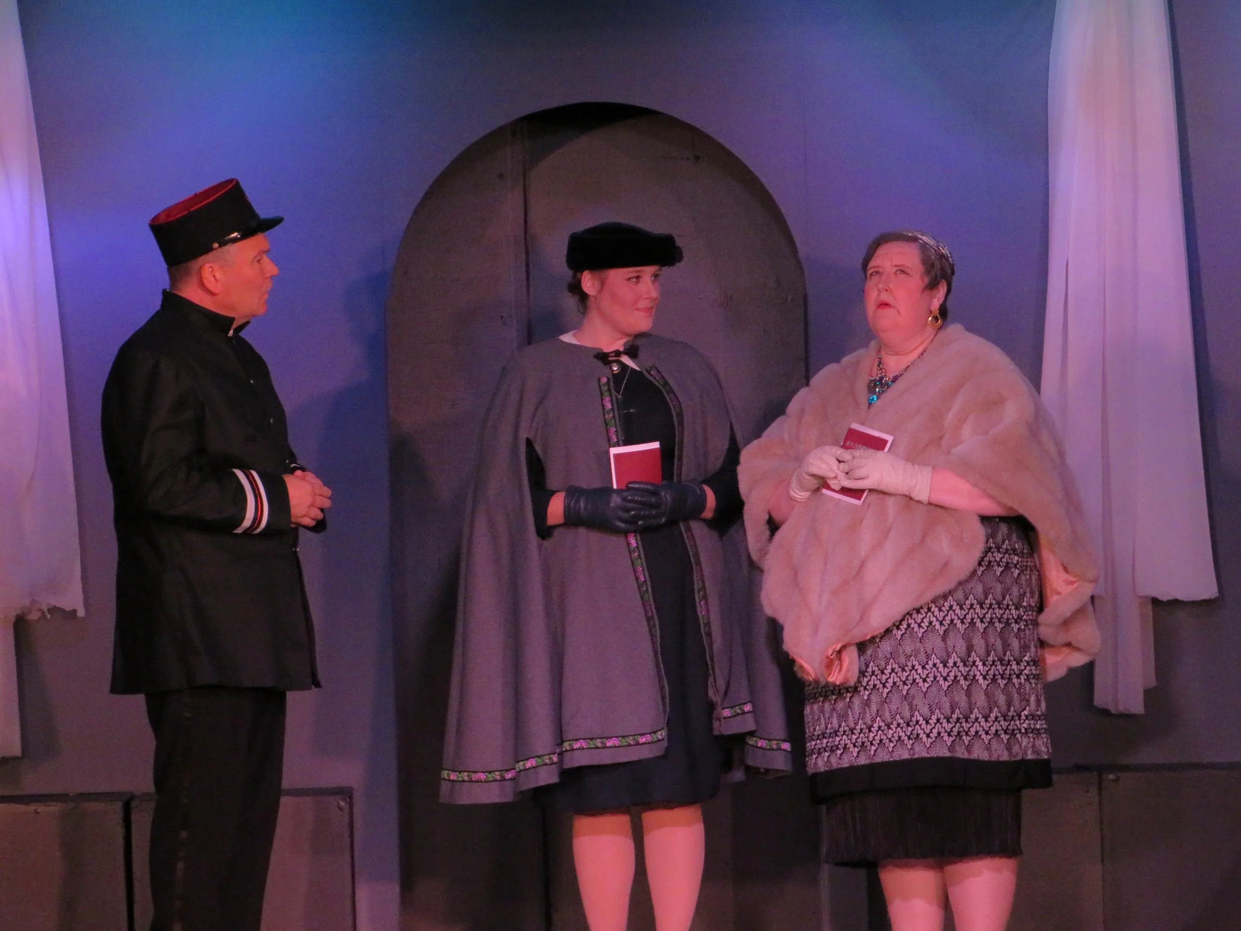 Three actors on stage in vintage costumes performing in a play. One man in uniform on the left, a woman in a gray cloak and black hat in the middle, and an older woman with short gray hair and a fur stole on the right.