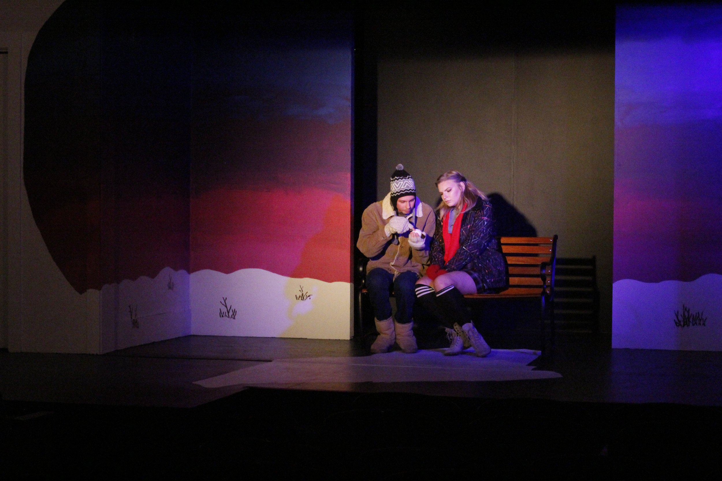 Two young women sitting on a bench on a stage with a painted snowy landscape backdrop, one wearing a winter coat and beanie, the other in a plaid jacket and thigh-high socks, both looking at an item in their hands.