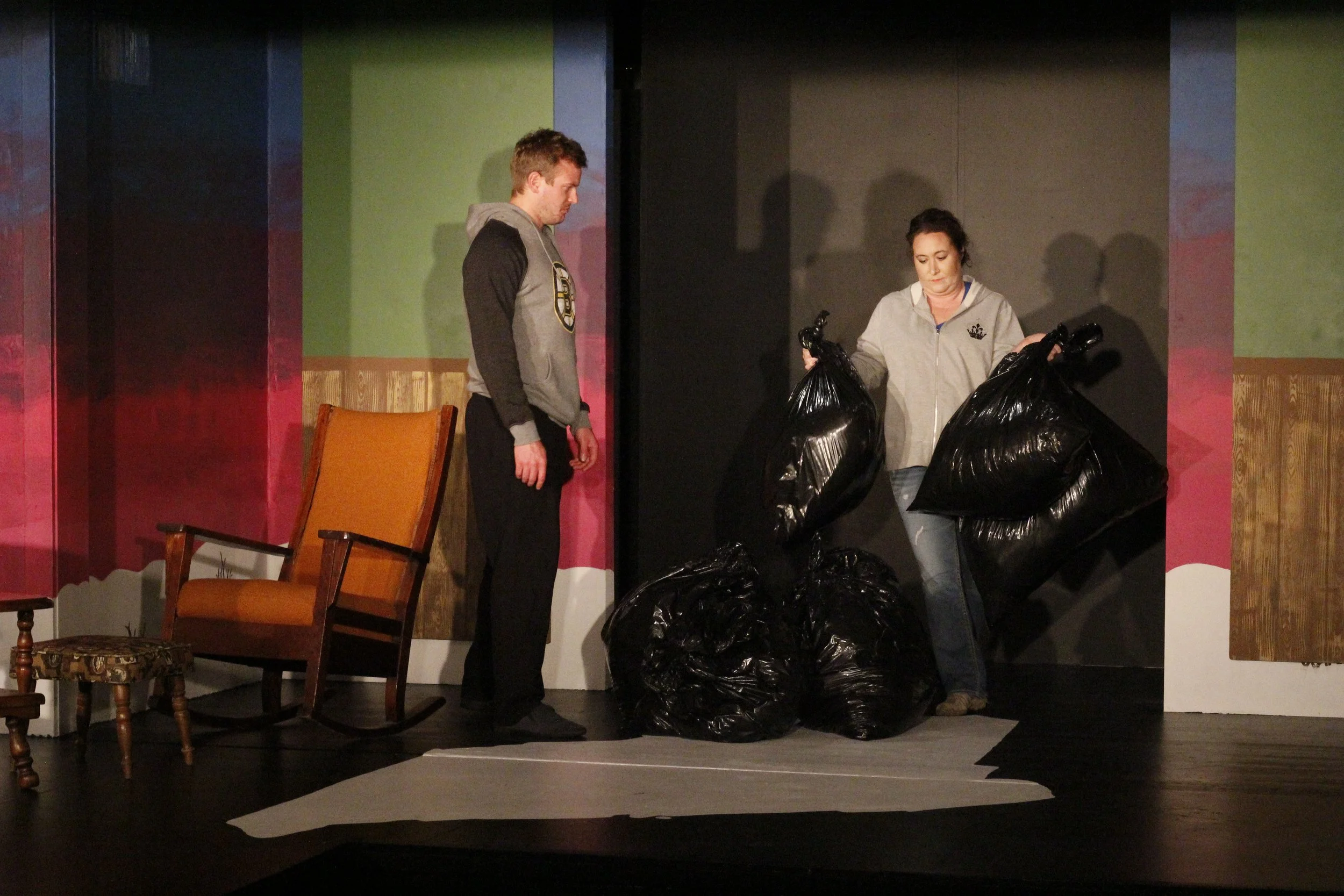 Two people, a man and a woman, standing on a stage with several large black garbage bags. The man is looking down at the bags, and the woman is holding two bags, seeming to inspect or organize them. The stage has a colorful painted backdrop and some 