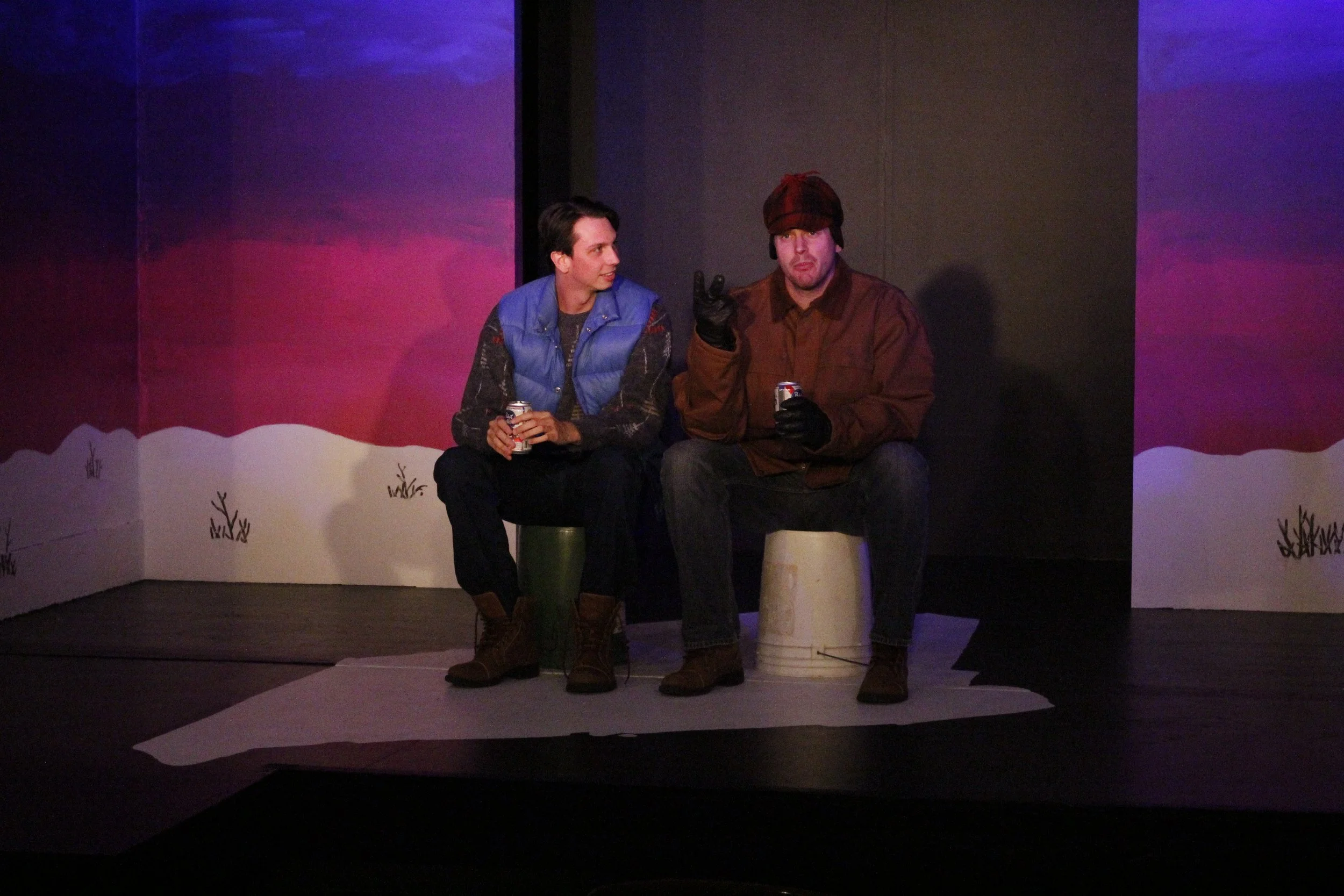Two men sitting on barrels on a stage, one wearing a blue vest and the other in a brown jacket, holding cans, with painted background resembling outdoors.