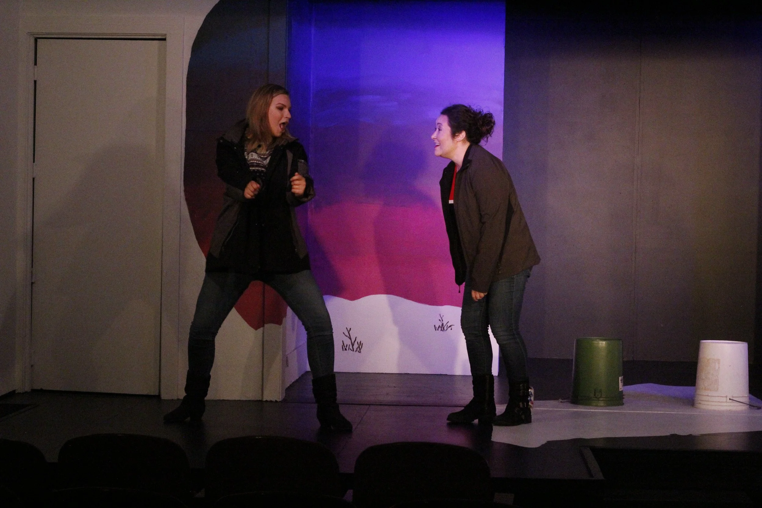 Two women are performing on a stage with a colorful background. One woman is holding a microphone and appears to be singing or speaking passionately. The other woman is facing her, smiling or laughing. There are three buckets on the stage floor behin