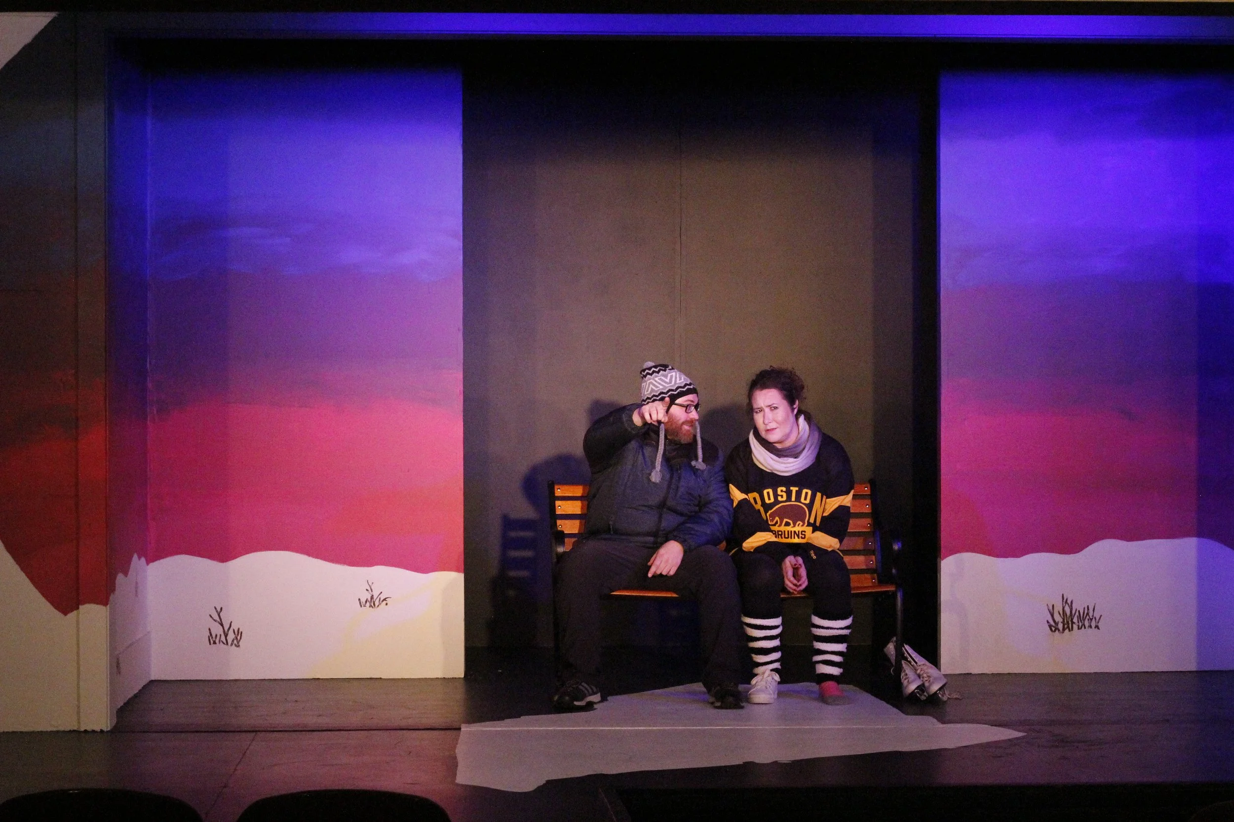 Two actors sitting on a bench on stage, one wearing a winter hat and the other wearing a Boston Bruins hockey jersey, with painted snowy mountain scenery background.