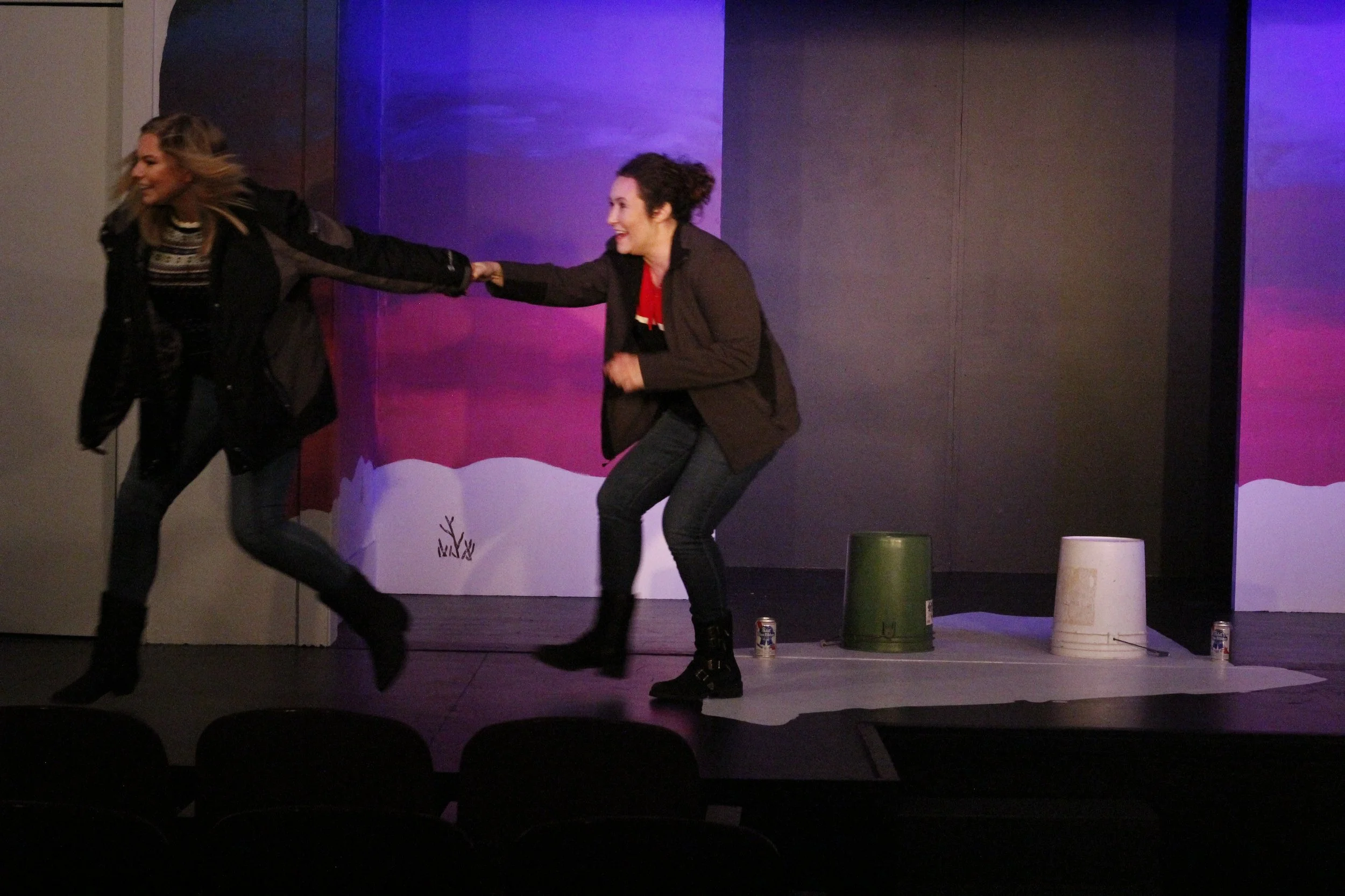 Two women are on a stage, one is pulling the other by the arm, both are smiling and appear to be pretending to run or chase each other, with a colorful abstract background behind them.