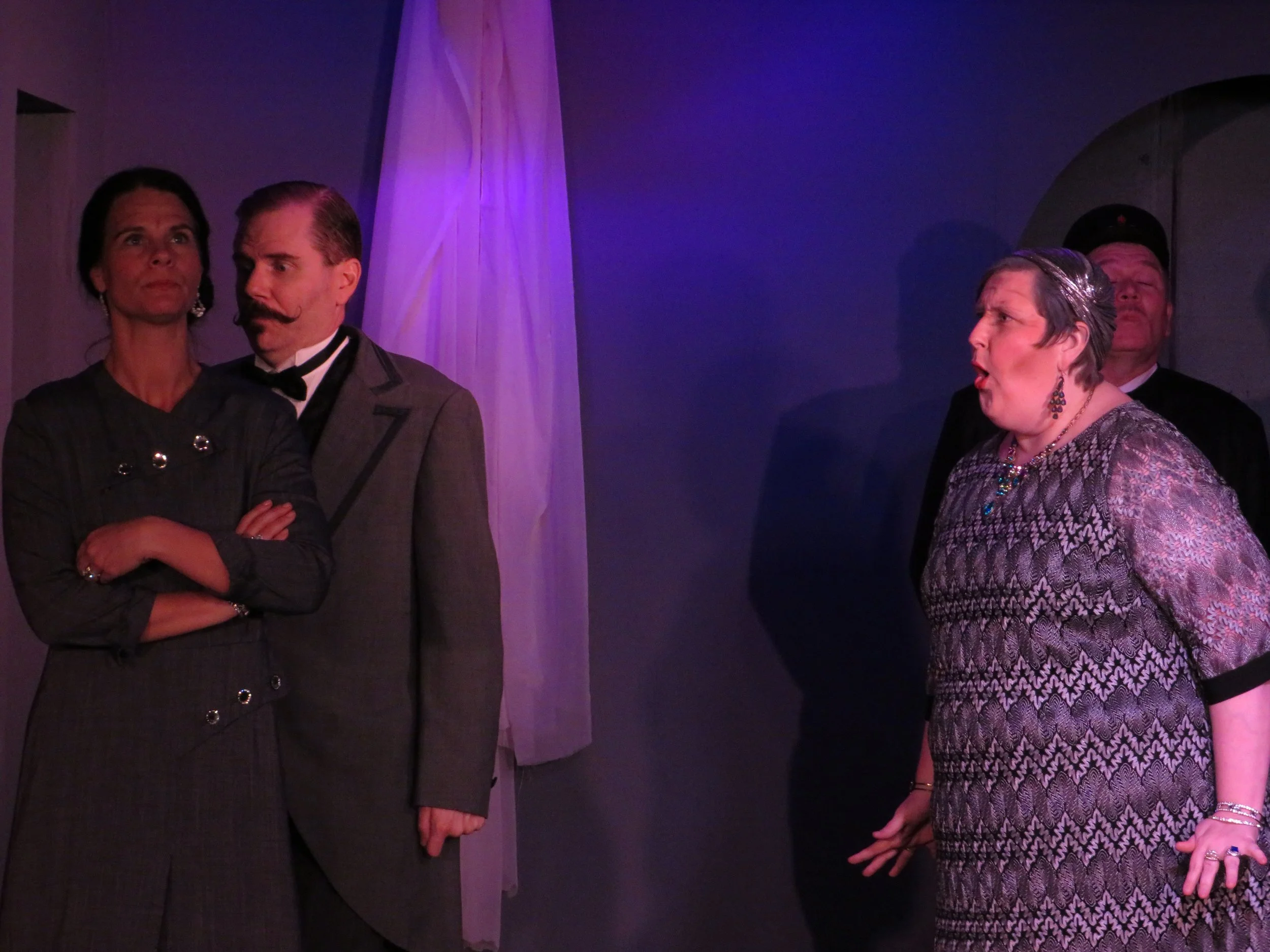 Three adults on stage, two women and one man, dressed in vintage or formal clothing, with two people in the background. The woman on the left has dark hair, the man in the middle has slicked back hair and a mustache, and the woman on the right has sh