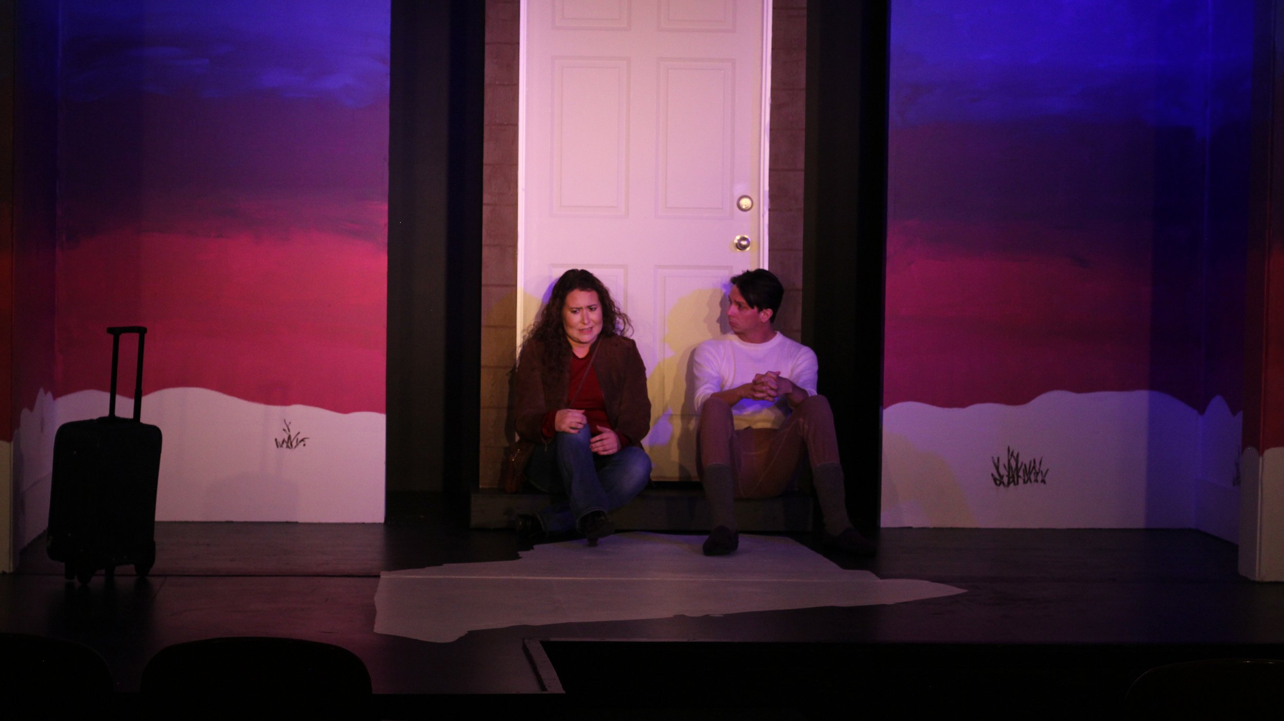 Two actors sitting on the stage, a woman with curly hair and a man, staged in front of a door with painted scenic backgrounds, a suitcase placed on the floor to the left.