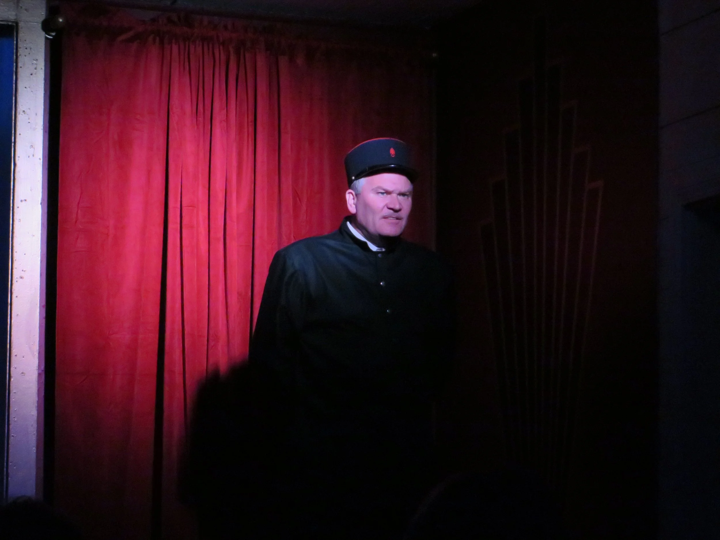 A man dressed as a traditional German or Bavarian performer stands in front of a red curtain, wearing a dark uniform with a hat.