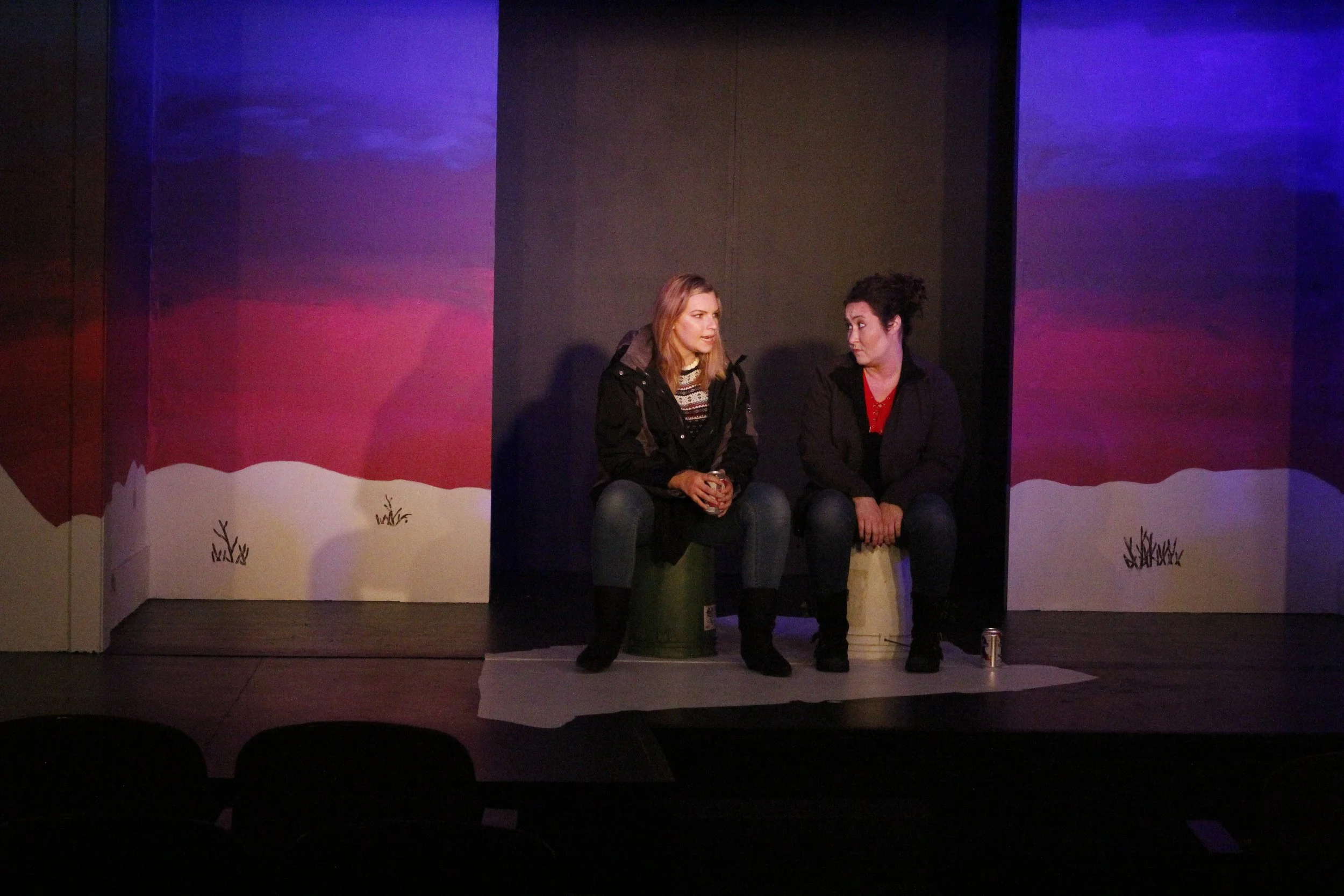 Two women sitting on stage with painted winter landscape background, having a conversation, with a soda can on the floor nearby.