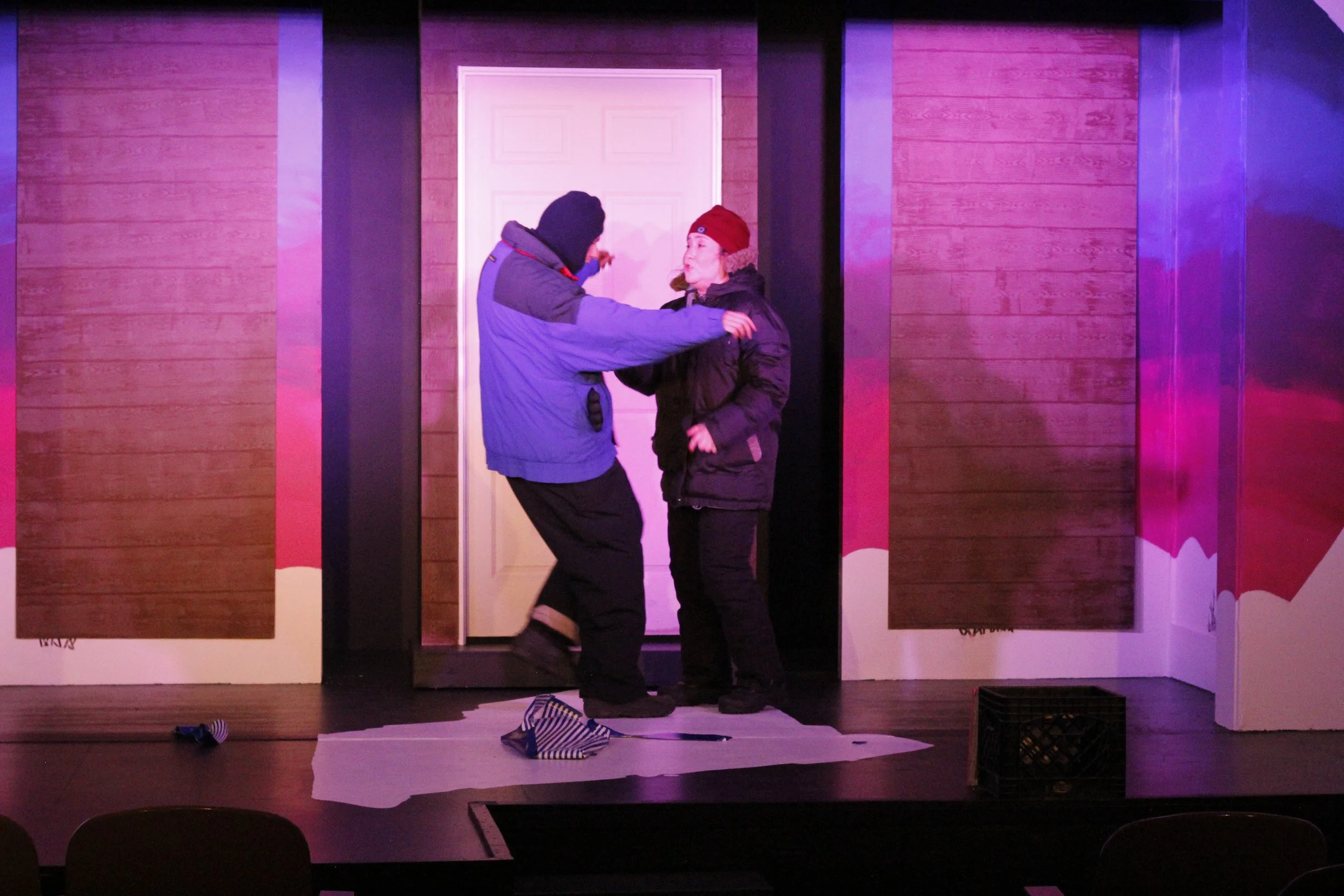 Two actors perform on stage in front of a wooden backdrop with a pink and purple lighting. One actor, wearing a blue jacket, faces another actor in a black jacket and red hat, engaged in a tense moment. Props including a fabric on the floor and a mil