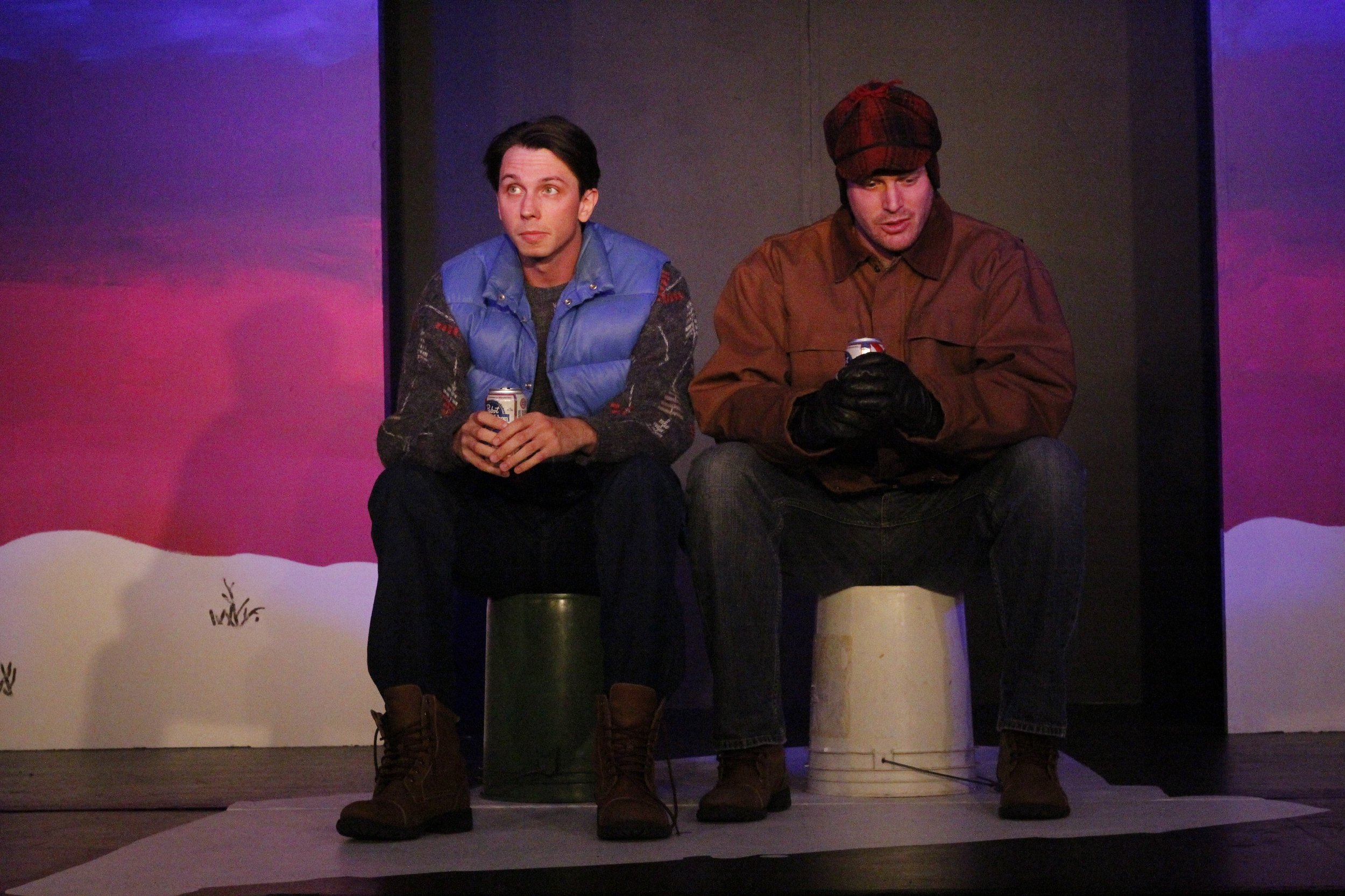Two men are sitting on buckets on a stage, each holding a can of beer. The man on the left is wearing a blue vest over a sweater, while the man on the right is dressed in a brown jacket with a red plaid hat. The background is painted with purple and 