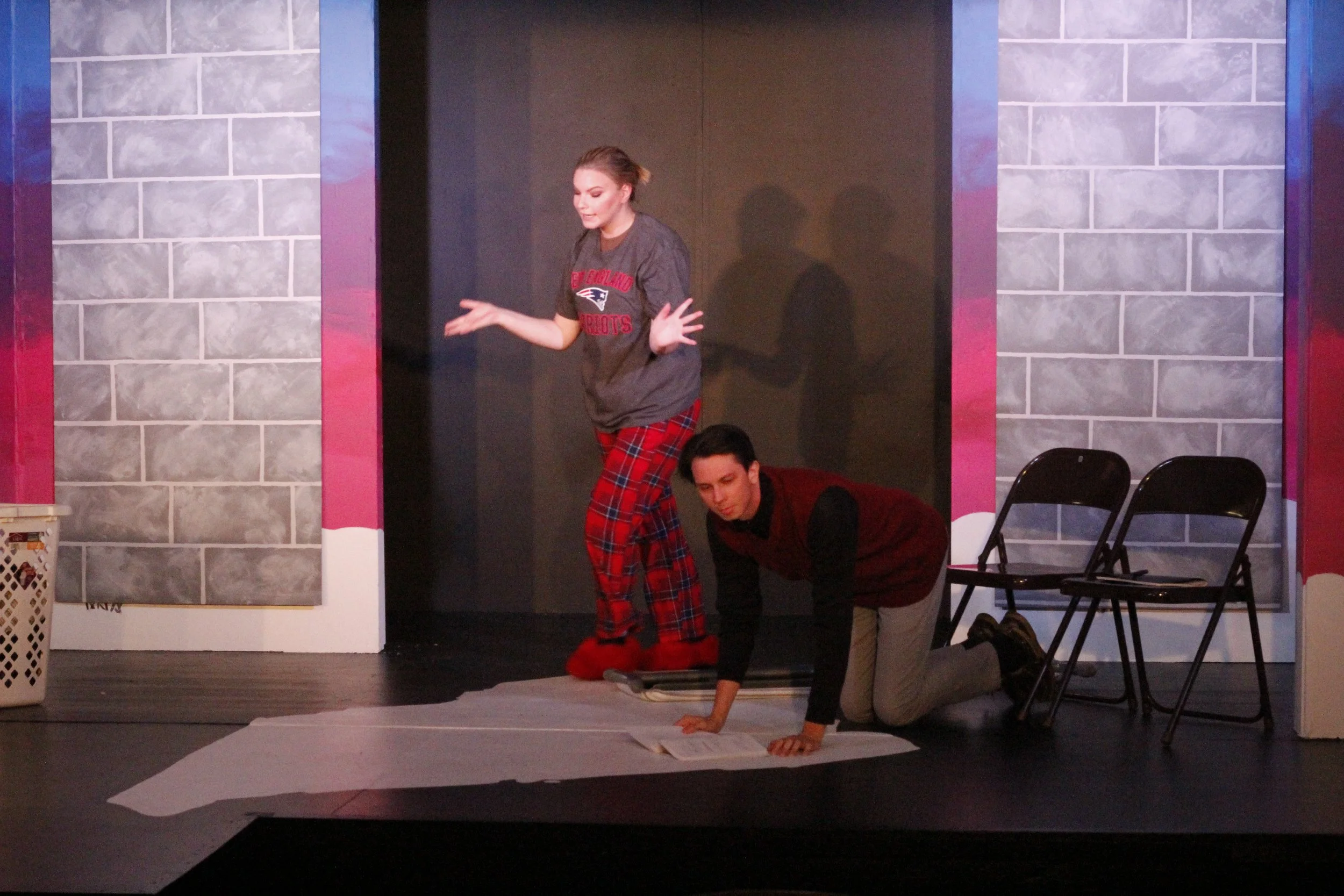 Two actors perform on stage; one is standing with pajamas and a T-shirt, the other is on all fours reading a script.