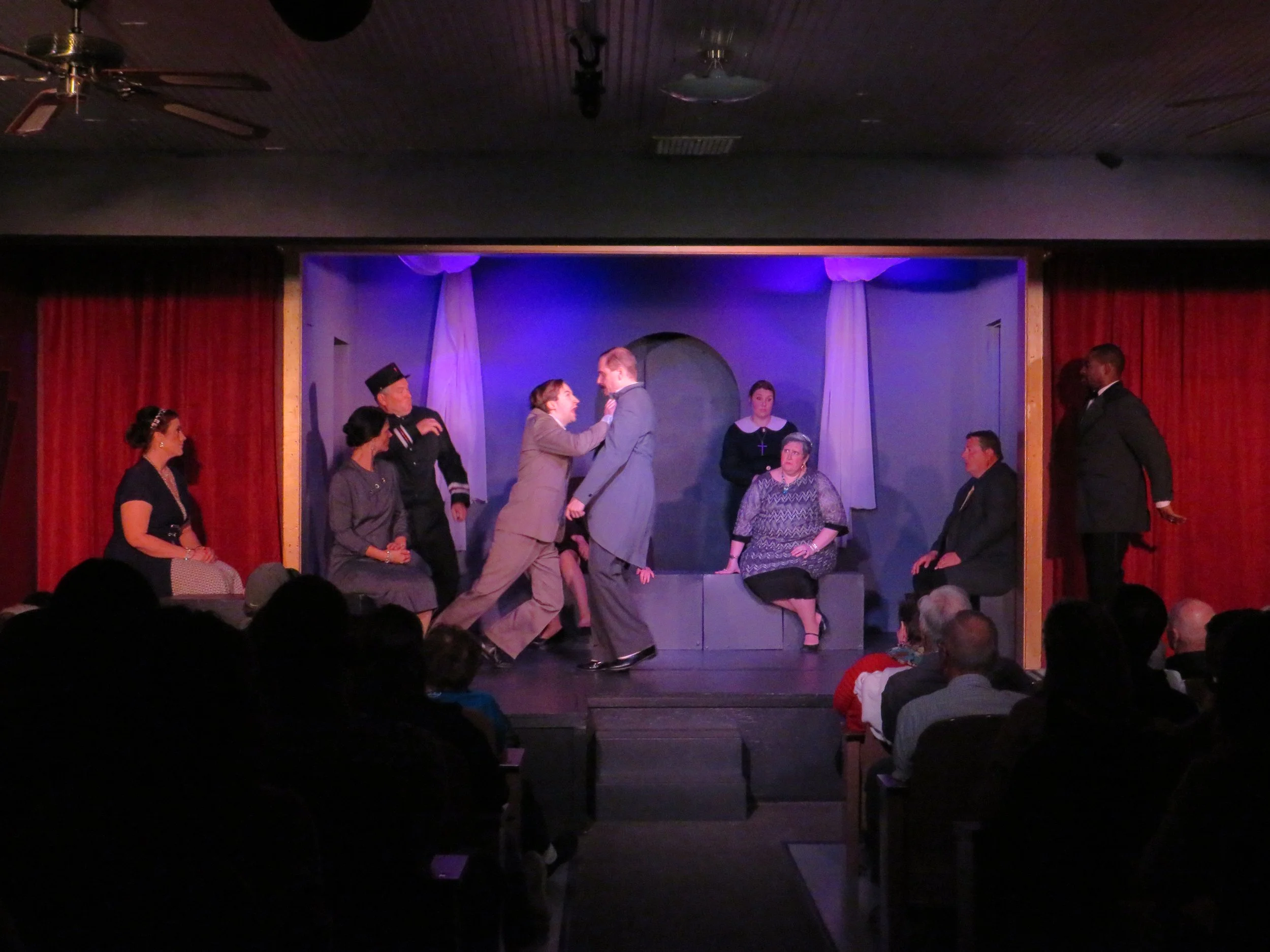 A theatrical stage with actors performing a scene, audience in foreground, red curtains on sides, blue backdrop, and overhead lighting.