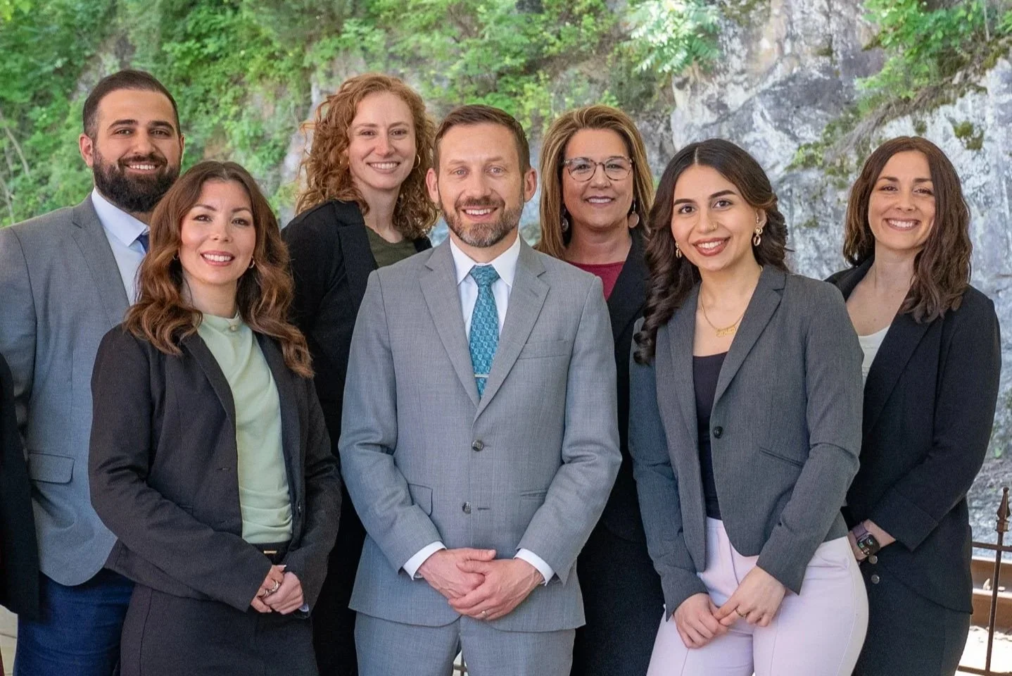 Group of eight diverse professionals, four women and four men, dressed in business attire, standing outdoors in front of a green, rocky landscape.