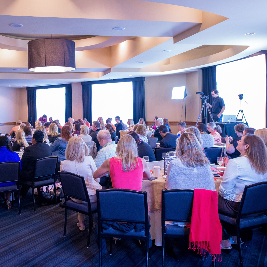 A large group of people seated at round tables in a conference room, attending a presentation or seminar, with cameras and lighting equipment set up at the front.