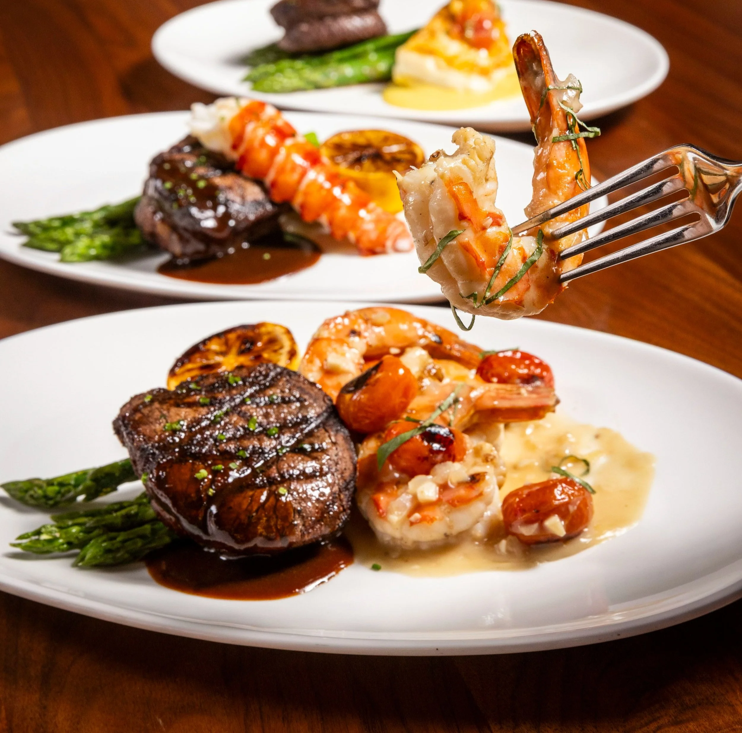 A grilled filet with shrimp on a fork and a grilled filet with a lobster tail behind it on a wood table.