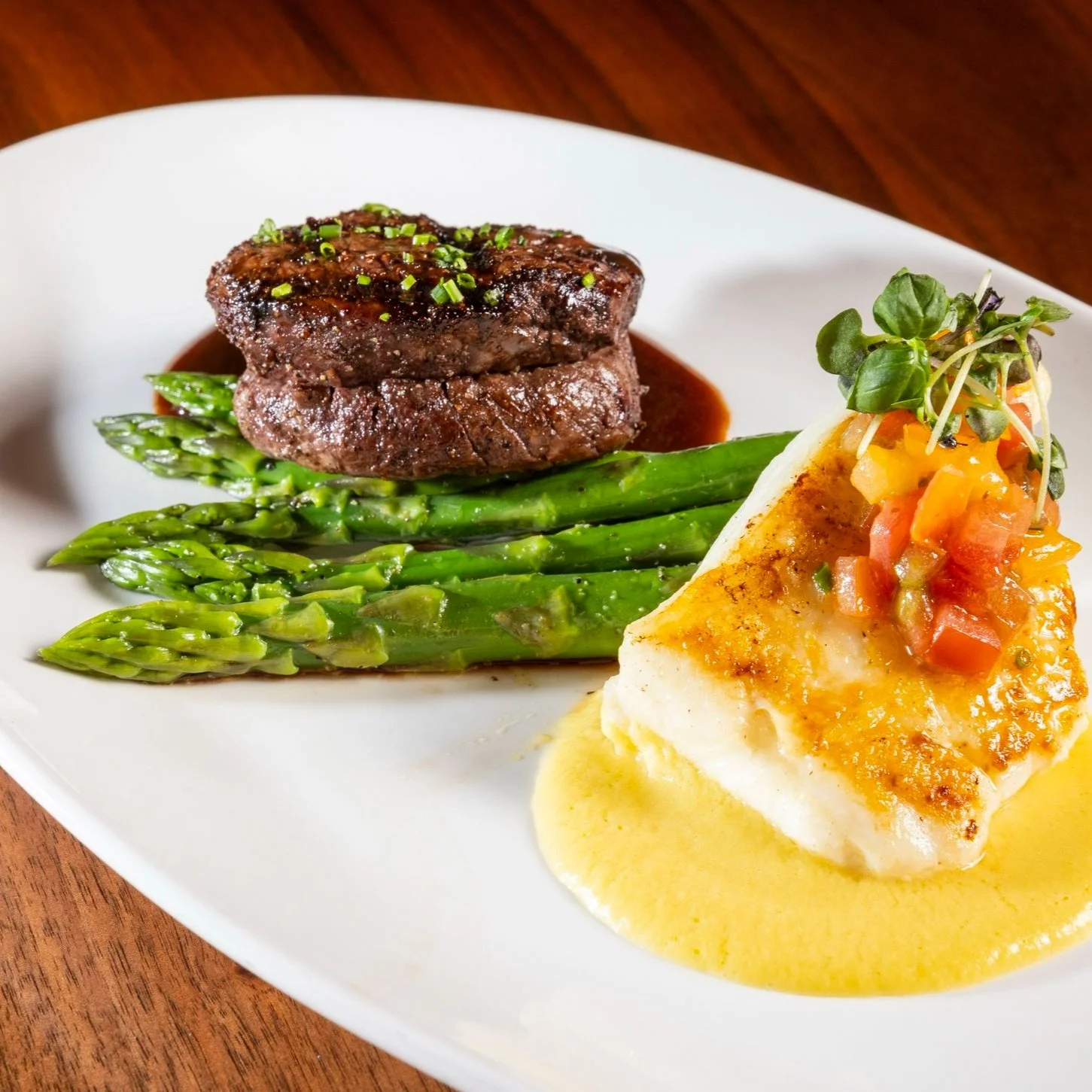 A plate with a filet topped with demi sauce, grilled asparagus spears, and a piece of halibut garnished with diced tomatoes, herbs, and a creamy yellow sauce.