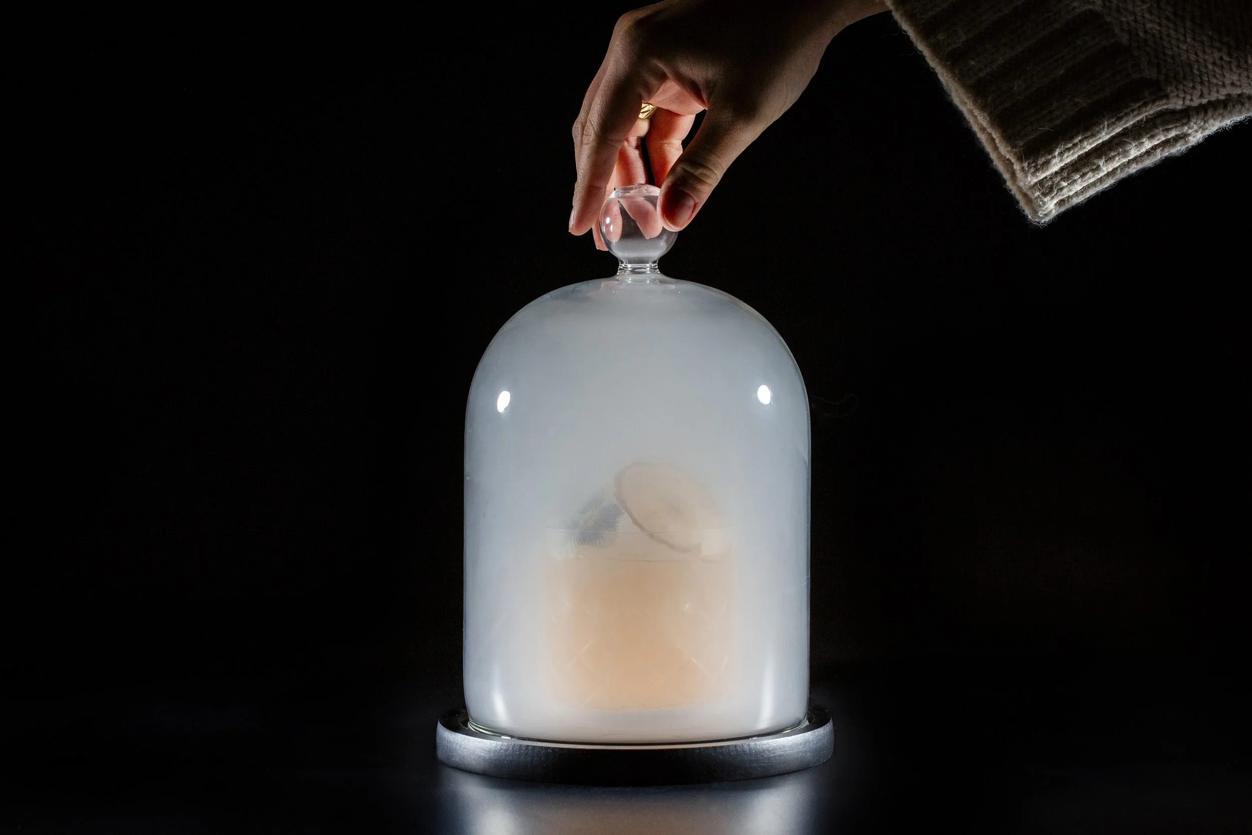 A hand removing the glass cover of a small glass dome with a foggy appearance, revealing a cocktail inside, on a black surface with a dark background at Alara in Cincinnati..