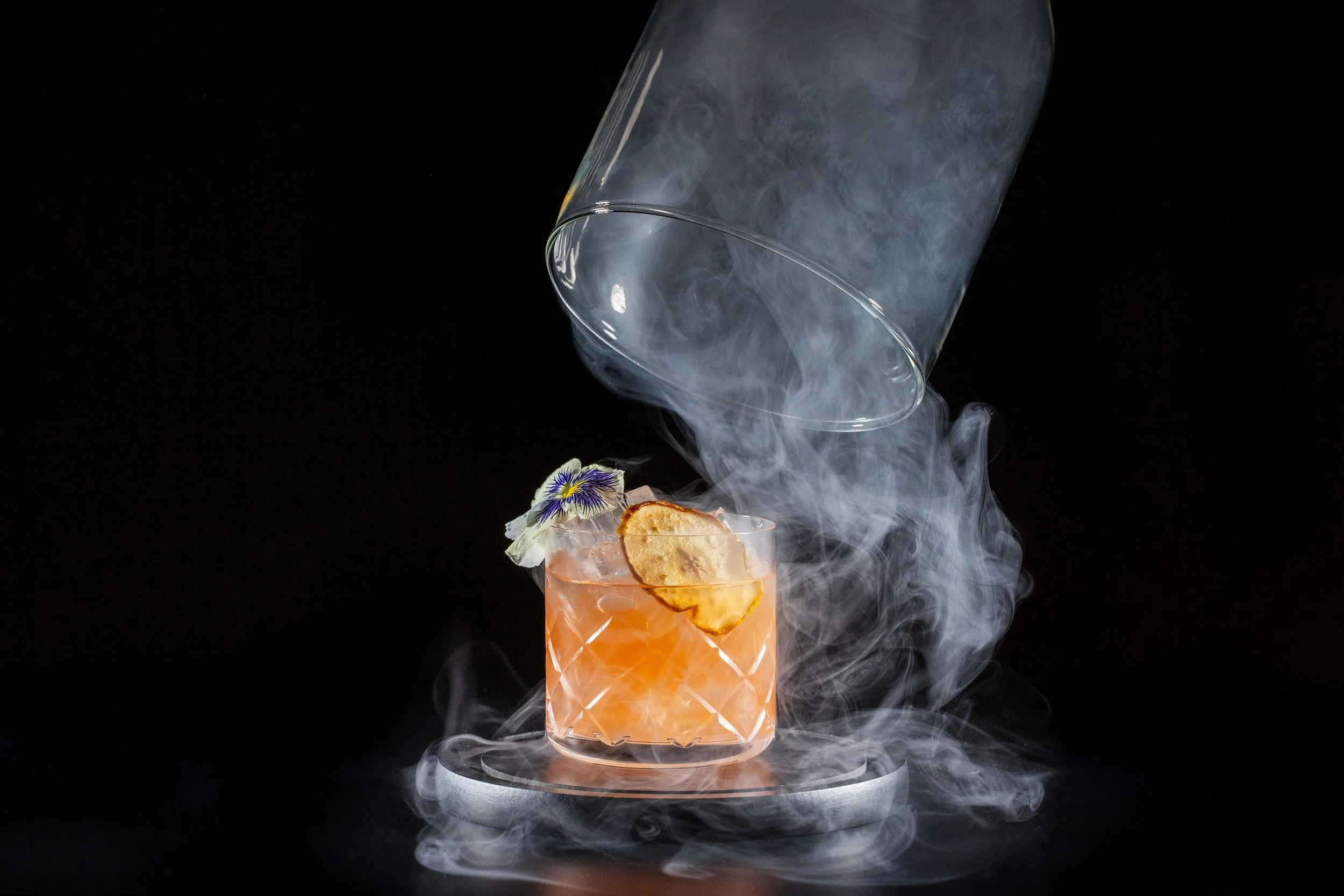 Alara in Cincinnati, A cocktail glass with an orange-colored drink, garnished with a dried lemon slice and a purple edible flower, surrounded by swirling smoke or vapor, with an overturned glass above pouring more smoke, against a black background.