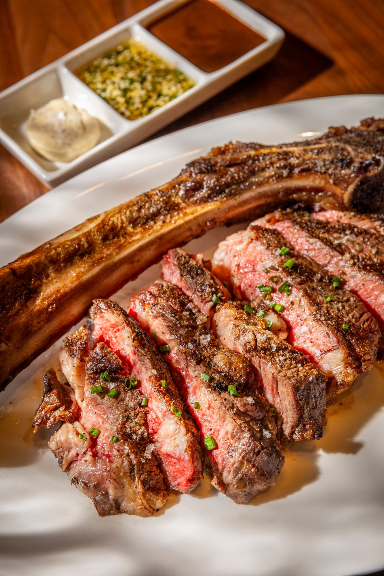 Alara in Cincinnati, Sliced Tomahawk steak with a bone, garnished with chopped green onions, served on a white plate, with side condiments in a compartmentalized dish in the background.