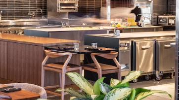 A view of the restaurant kitchen and a chef's table from the dining room with a green plant in the forefront