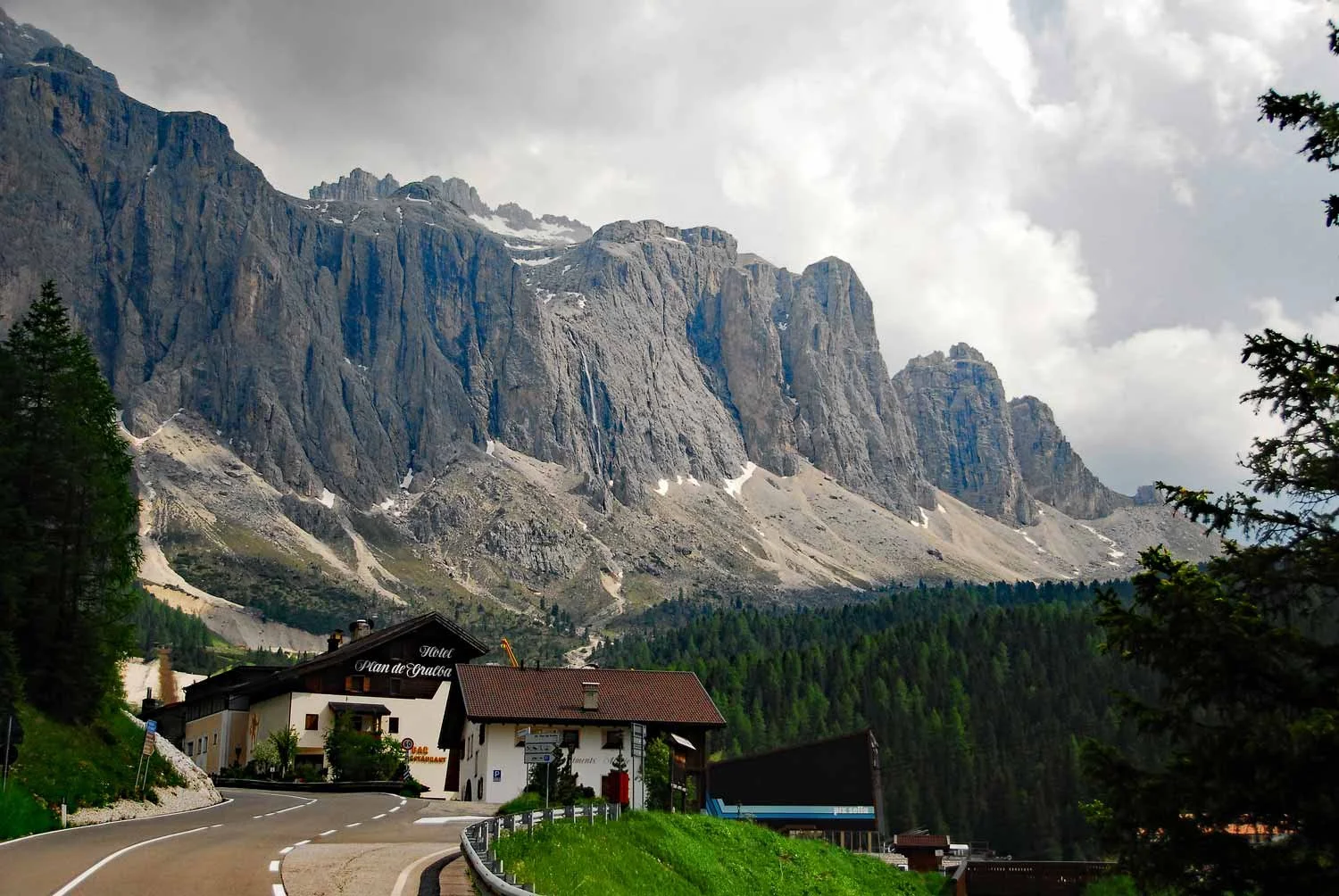 Mountain landscape with steep cliffs and rock formations, a forested area at the base, and a winding road with houses in the foreground.