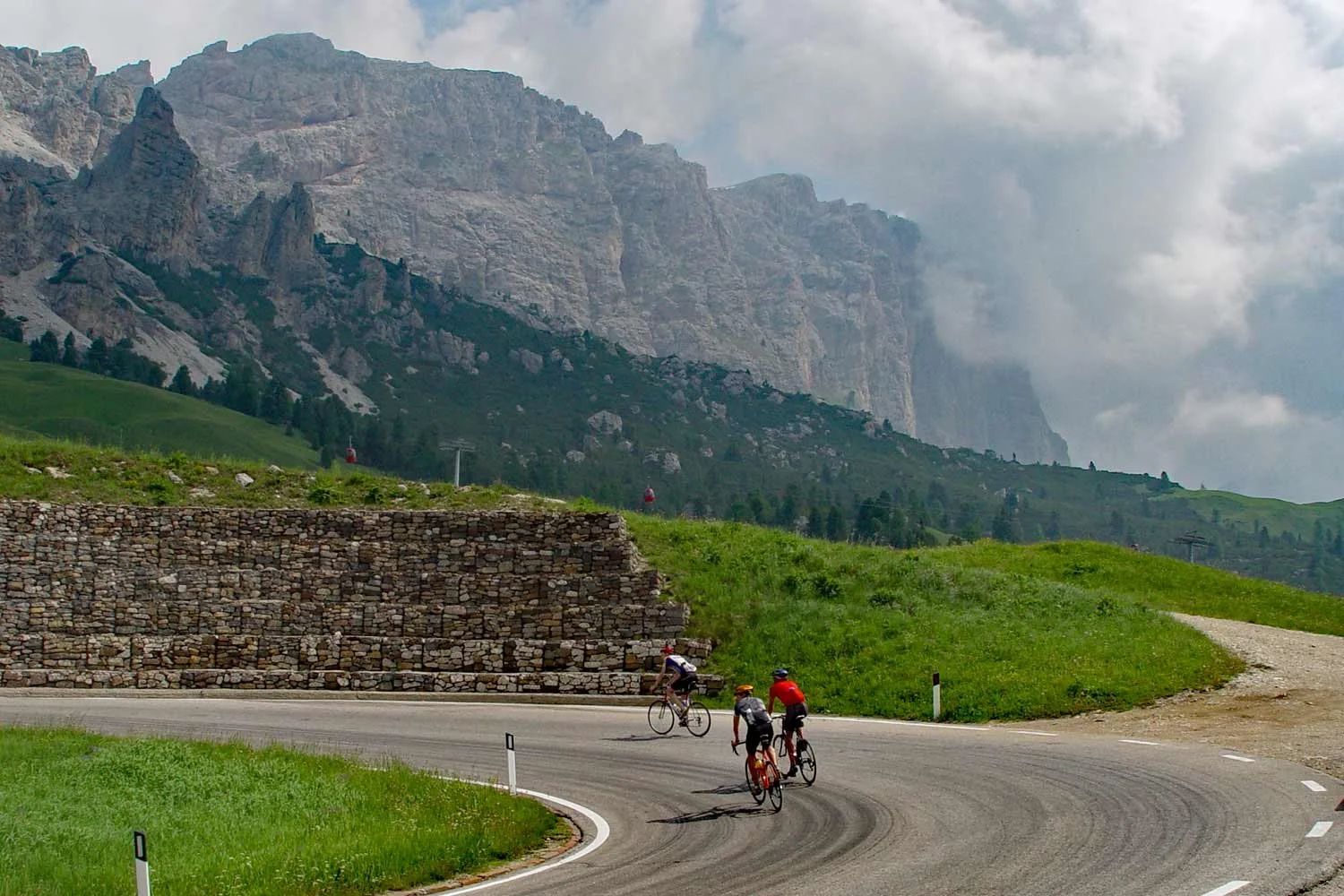 Three cyclists riding on a winding mountain road with a stone retaining wall on one side and green hills in the background, with gray cloudy sky and towering rocky mountains in the distance.