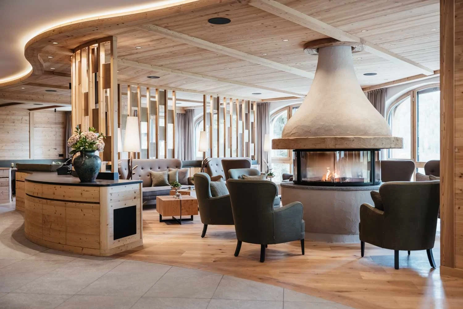 A cozy hotel lobby with a central fireplace, surrounded by armchairs and sofas, wooden accents, large windows, and a flower arrangement on a curved wooden counter.